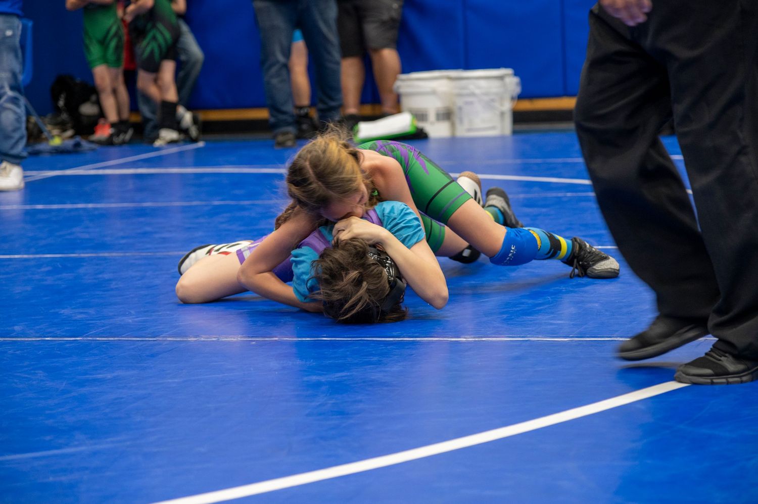 Two young children wrestling on a blue mat during a youth wrestling competition.
