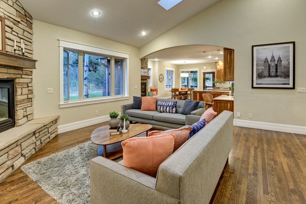 Cozy living room with stone fireplace, gray sofas, hardwood floors, skylight, and open kitchen with arched doorway.