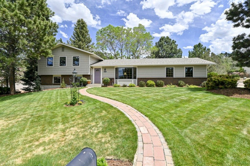 Ranch-style home with lush green lawn, mature trees, brick pathway, and well-maintained landscaping on sunny day.