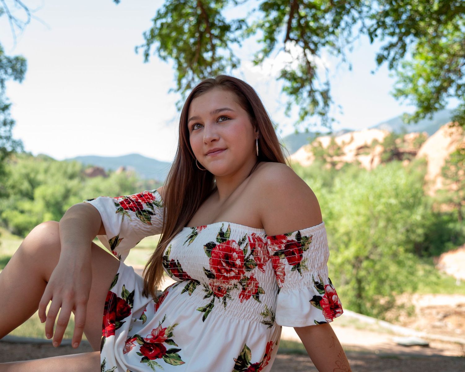 Young woman in floral off-shoulder dress posing outdoors with red rock mountains and green trees behind her.