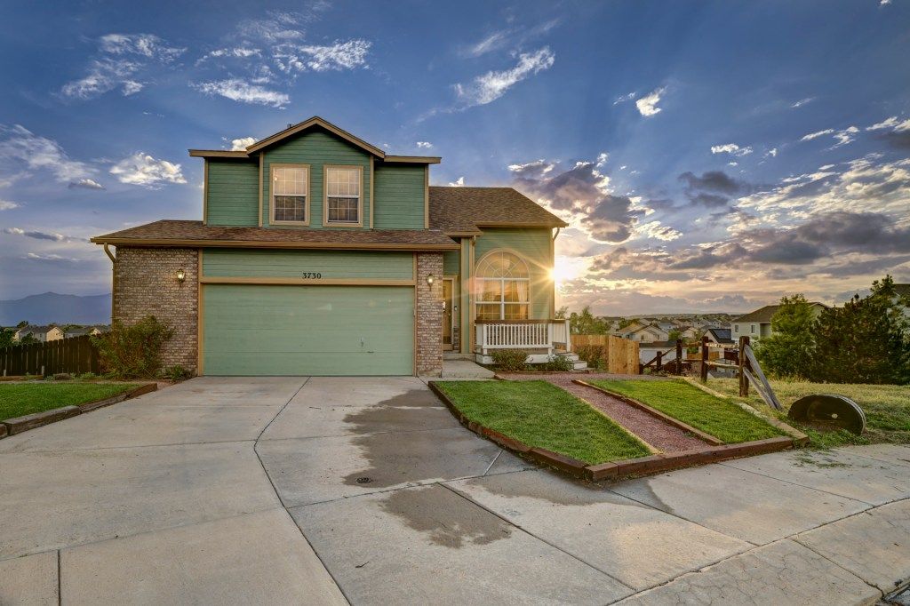 Two-story green and brick home with attached garage at sunset, featuring a concrete driveway and landscaped yard.