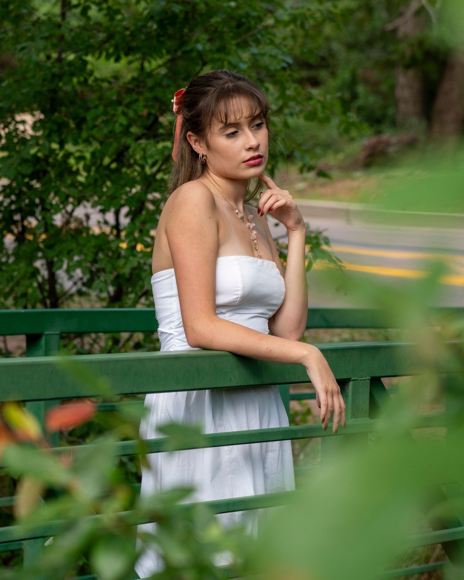 Young woman in white strapless dress posing thoughtfully on a green bridge surrounded by lush greenery.