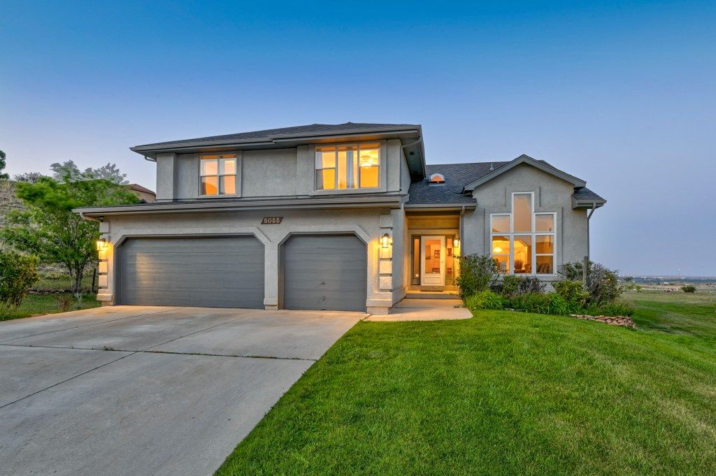 Two-story gray suburban home with three-car garage, lush green lawn, and warm interior lighting at dusk.