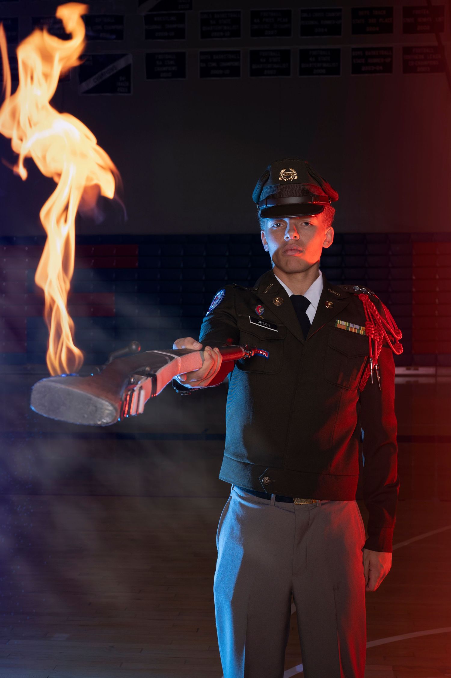 Military cadet in uniform holds a flaming torch in a dramatic dark indoor setting with red and blue lighting.