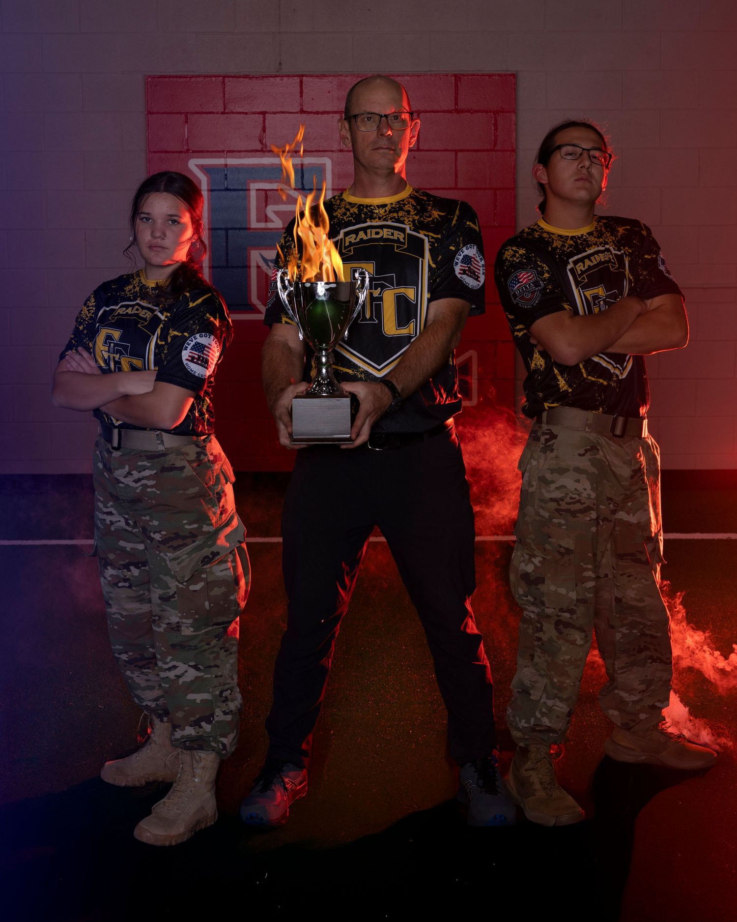Three JROTC Raiders members posing with a trophy against a dramatic fire-lit brick wall.