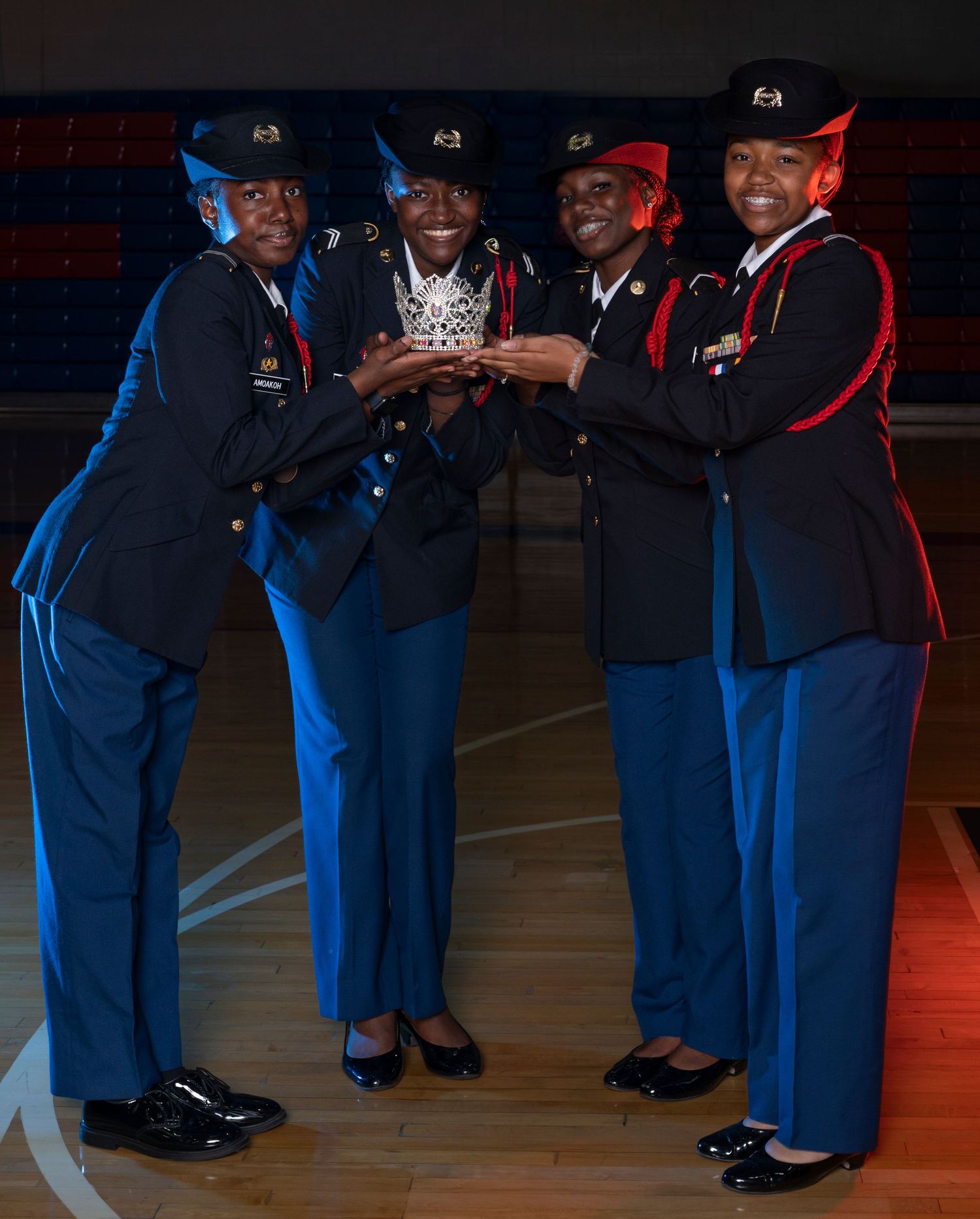 Four cadets in military dress uniforms proudly hold a trophy crown together on a gymnasium floor.