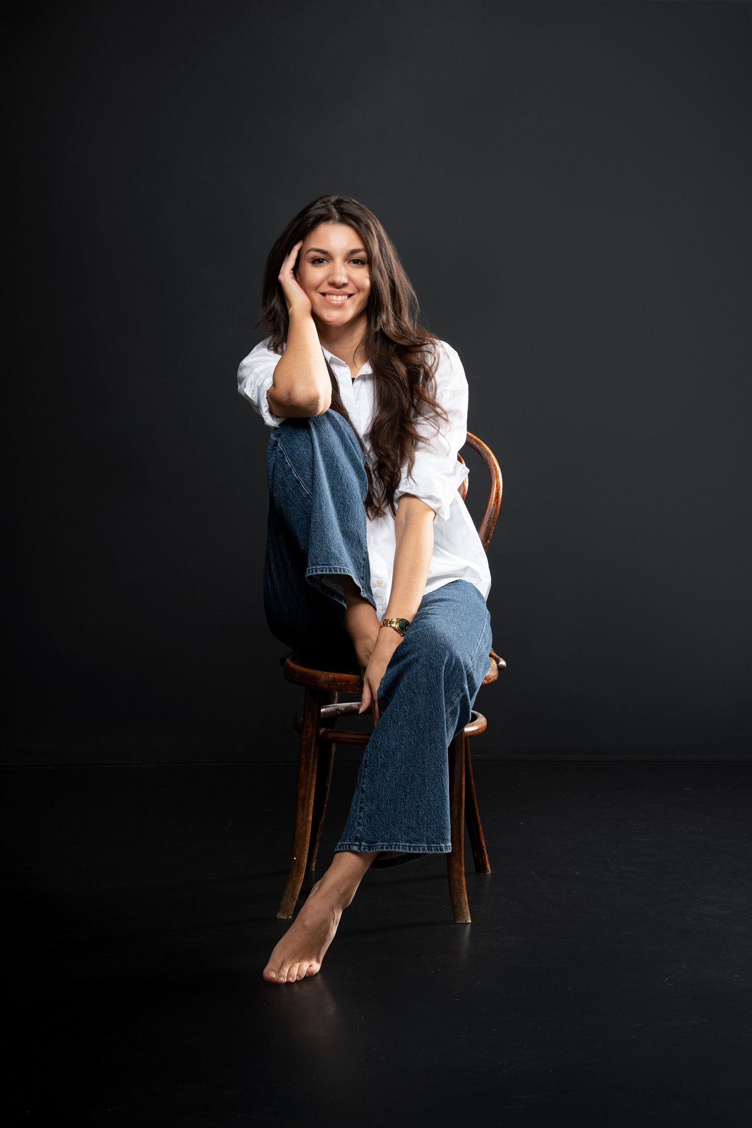Smiling woman in white shirt and blue jeans sitting barefoot on wooden chair against dark background.