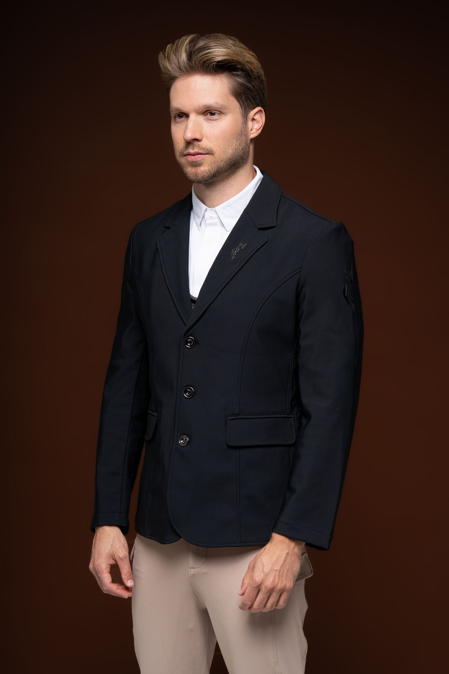 Confident man in navy blazer and beige trousers posing against a dark brown studio background.