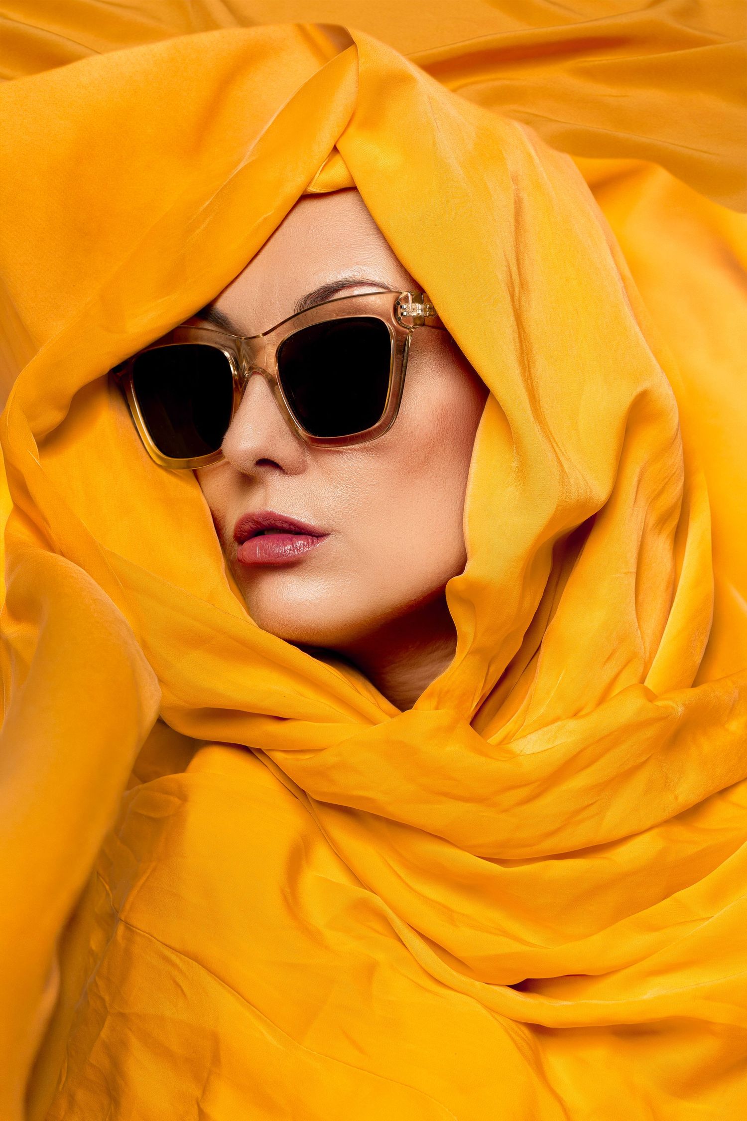 Woman wearing large square sunglasses wrapped in vibrant yellow fabric scarf on yellow background.