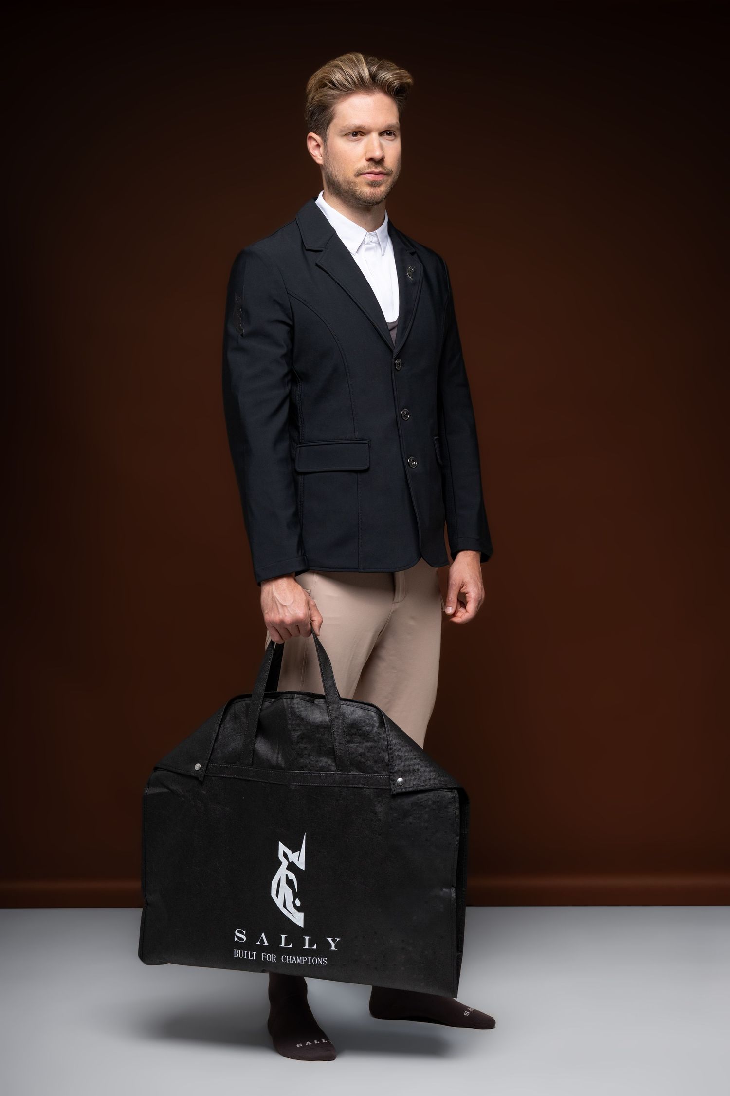 Man in navy blazer holding a black Sally equestrian gear bag against a dark brown background.
