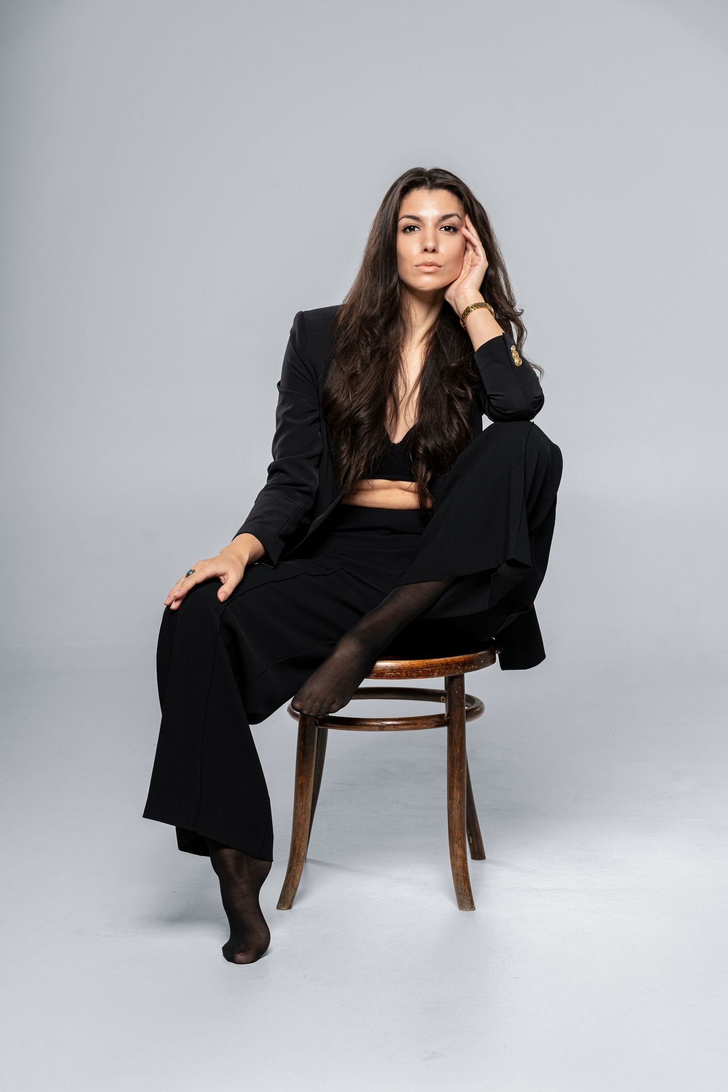 Brunette woman in black blazer and wide-leg pants sitting elegantly on wooden chair on gray background.