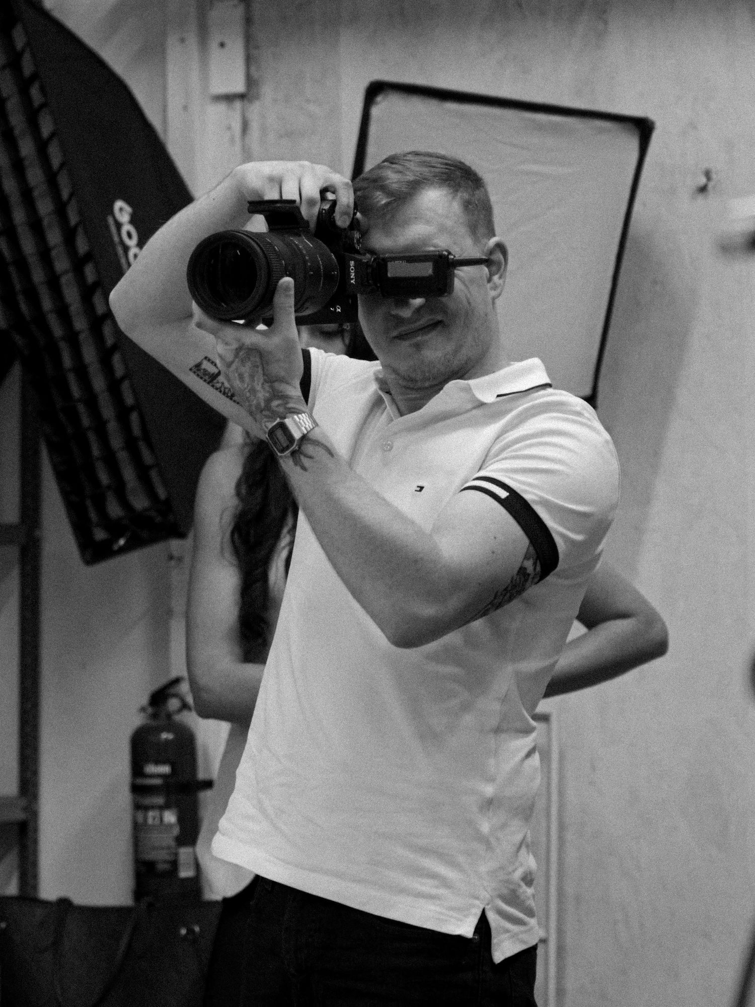 Black and white photo of a muscular man in a polo shirt holding a DSLR camera up to his eye in a photography studio.