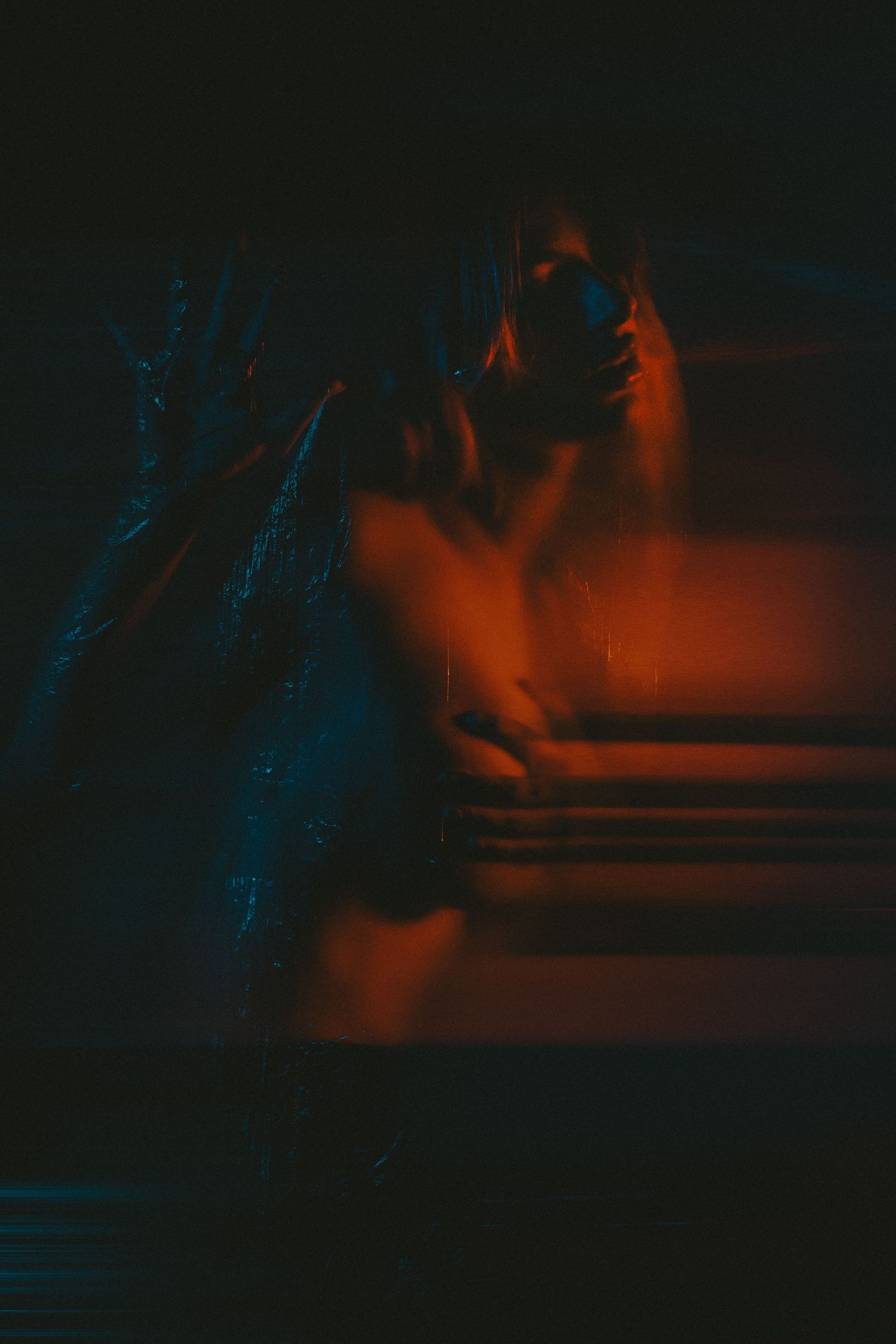 Dark artistic image of a figure wrapped in plastic illuminated by orange and blue neon light.