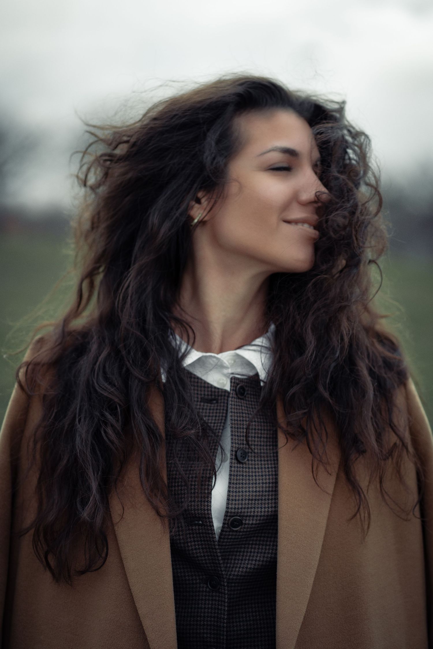 A woman with long curly dark hair blowing in the wind, smiling with eyes closed, wearing a brown coat outdoors.