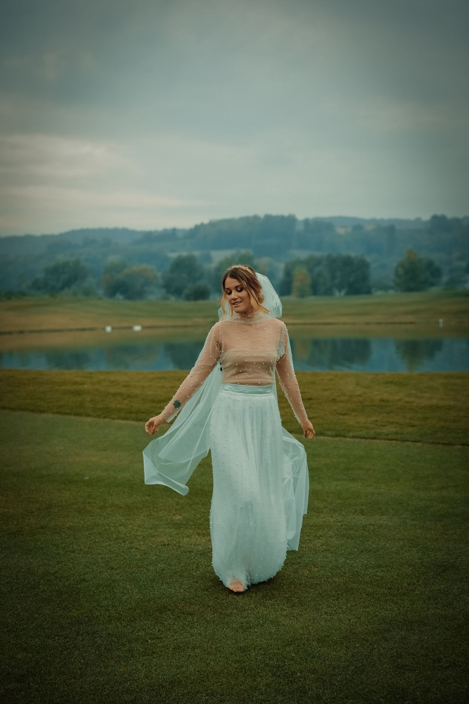 A bride in a light blue flowing skirt and veil walks gracefully on a green golf course near water.