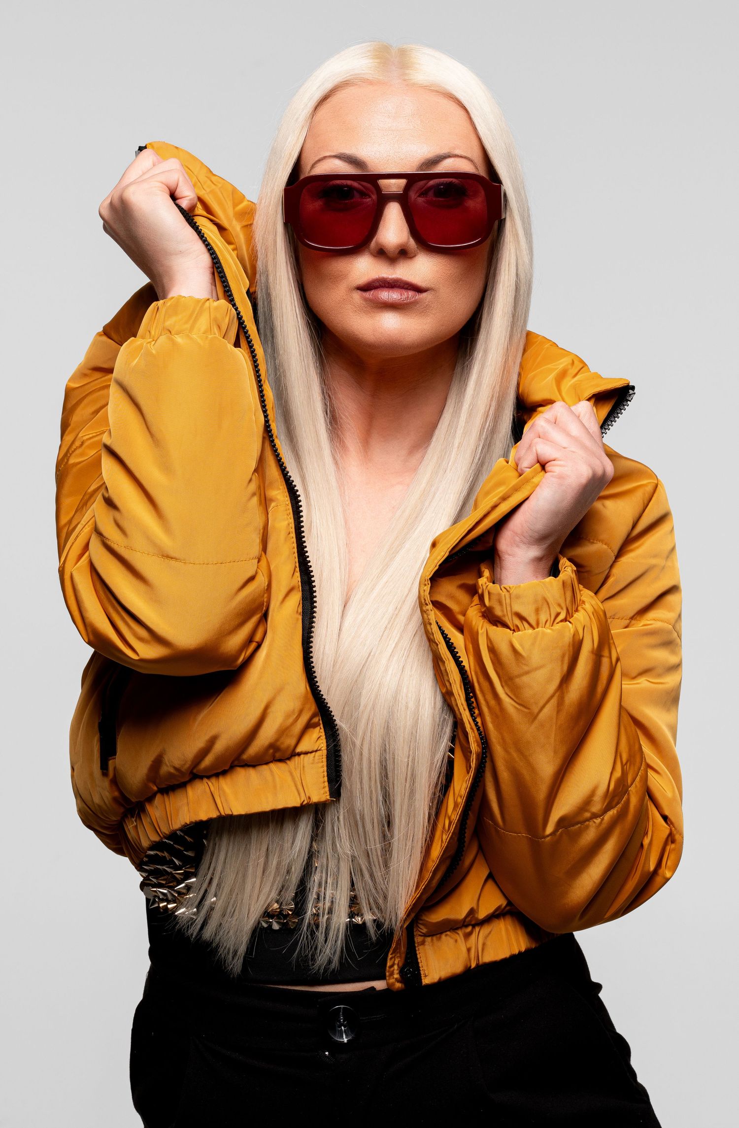 Blonde woman in yellow puffer jacket and red aviator sunglasses posing against a light gray background.
