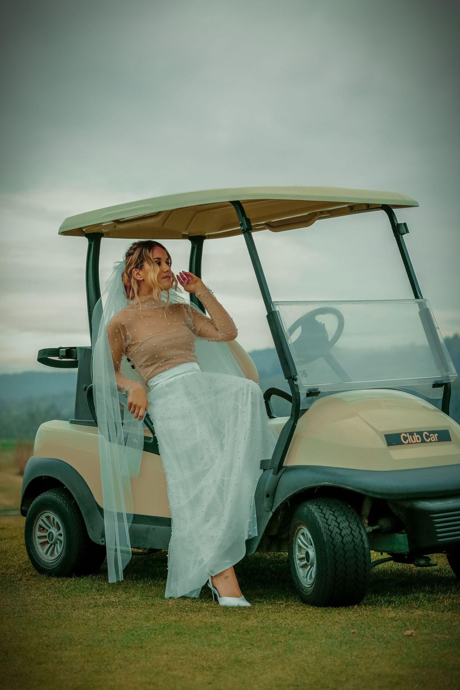 A bride in a light blue skirt and veil sits on a Club Car golf cart on an overcast day.