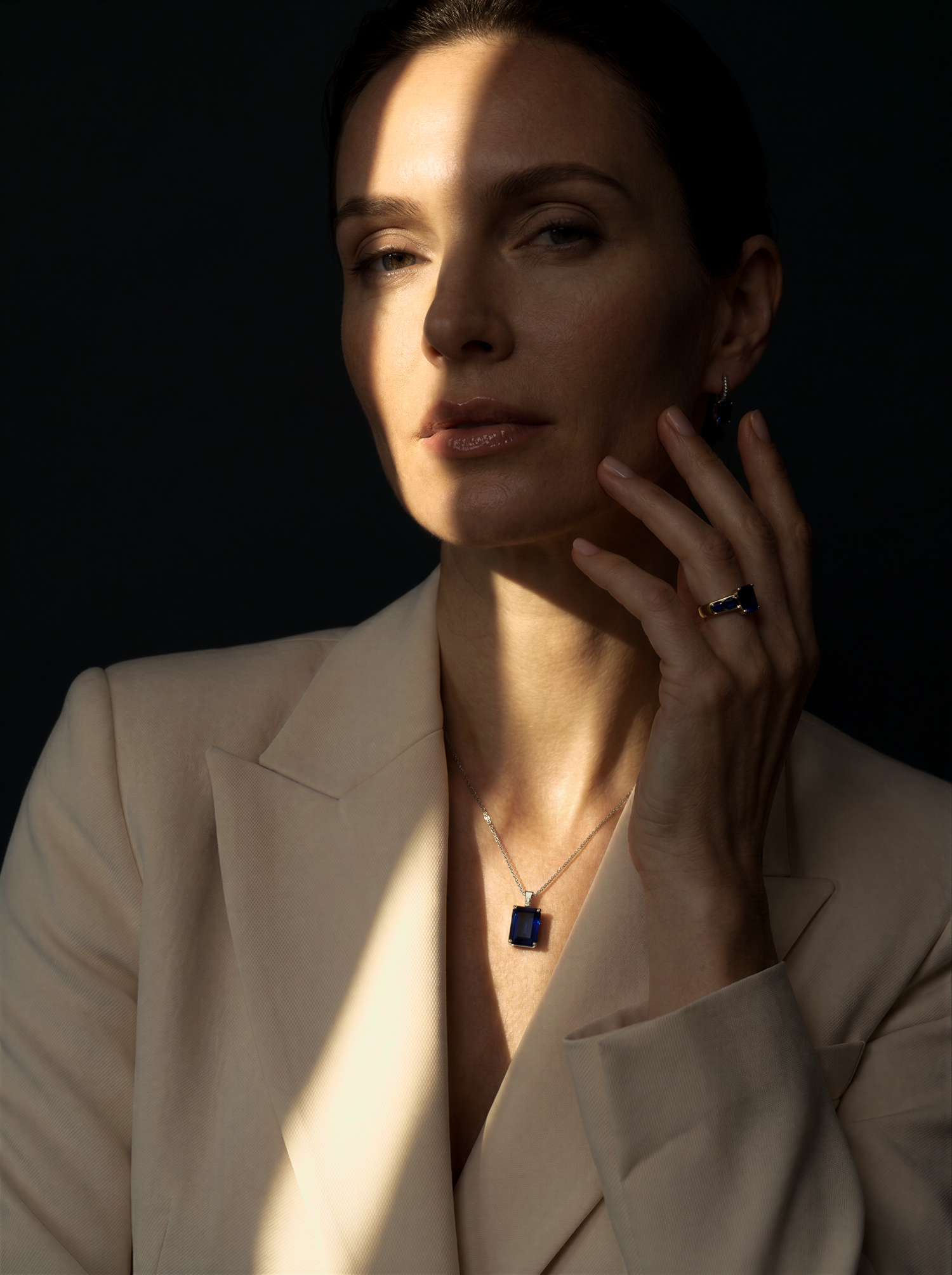 Elegant woman in beige blazer wearing a sapphire pendant necklace and ring, posed in dramatic studio lighting.