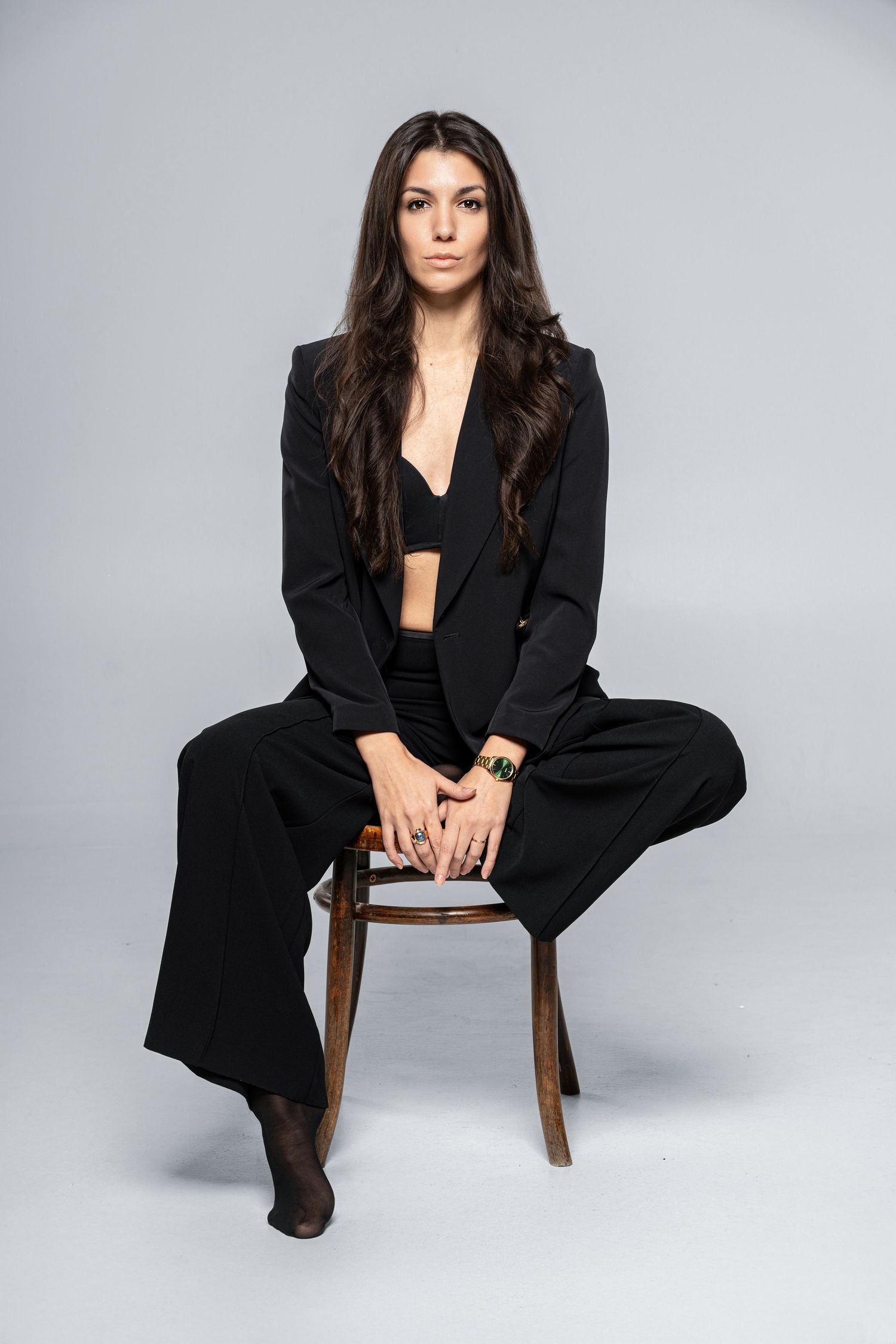 Confident woman in black suit with wide-leg trousers seated on wooden chair against gray studio backdrop.