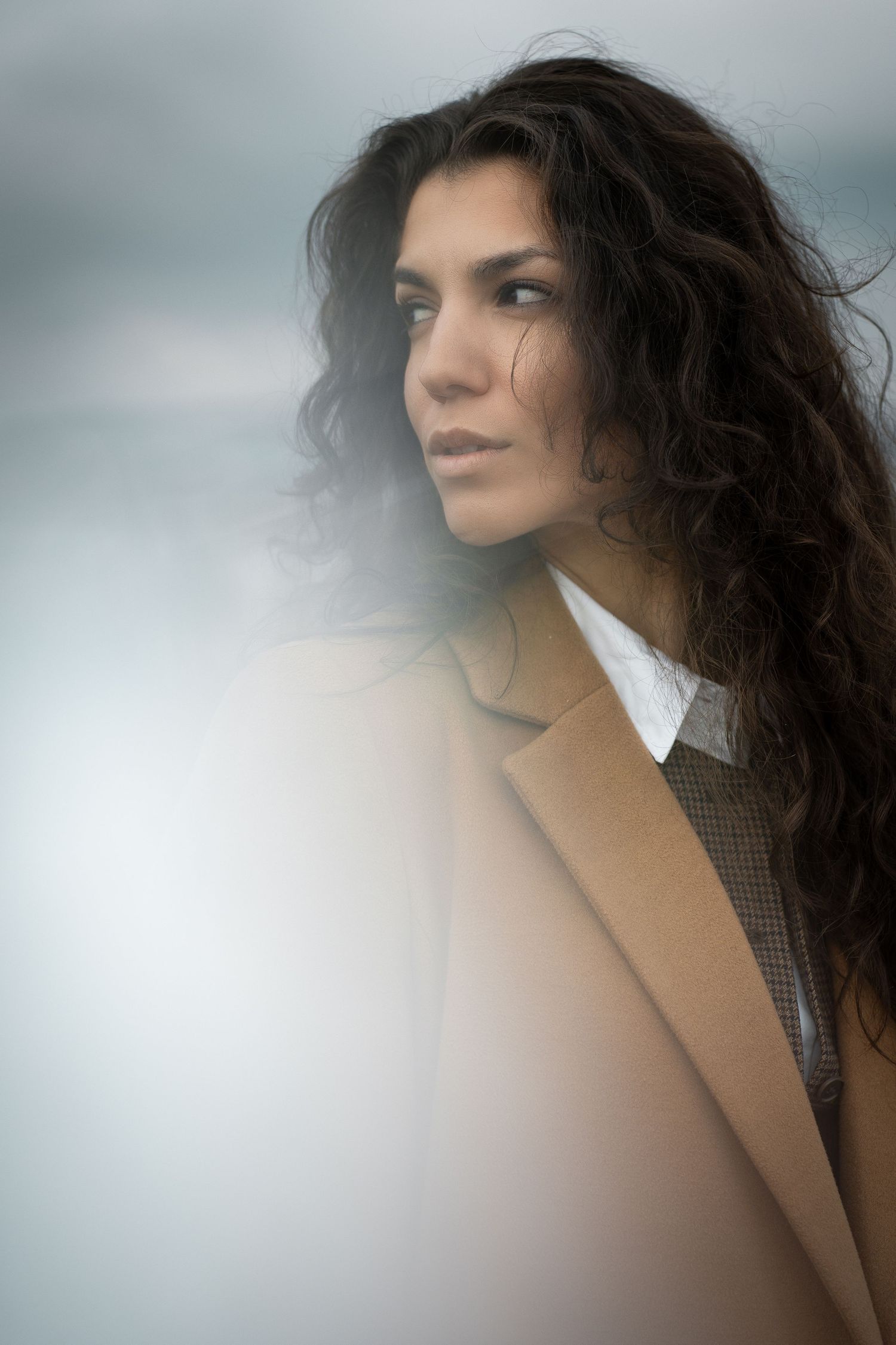 A woman with curly dark hair gazes into the distance, wearing a brown coat in a moody, foggy outdoor setting.