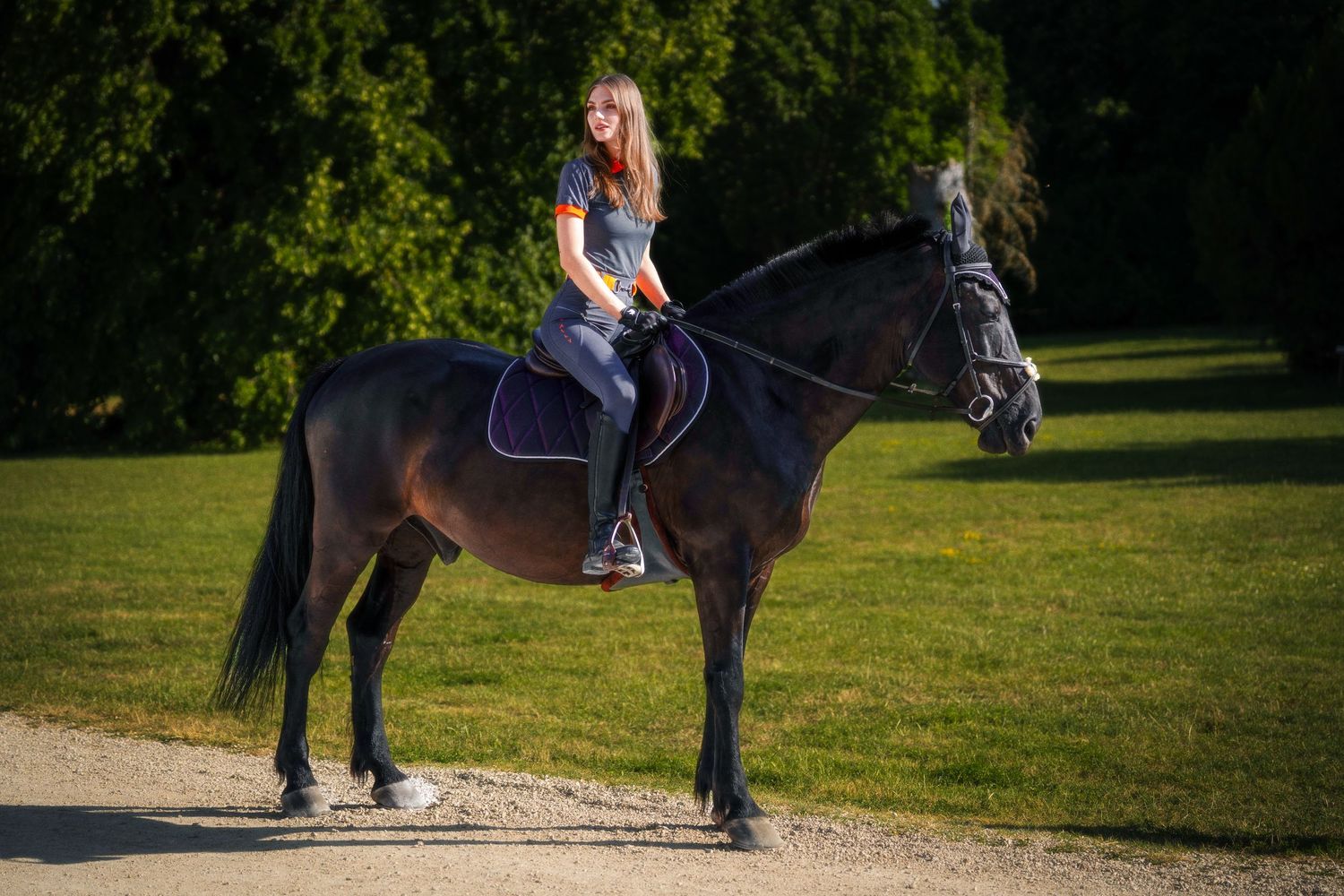 A young woman with blonde hair rides a black horse on a gravel path surrounded by lush green trees.