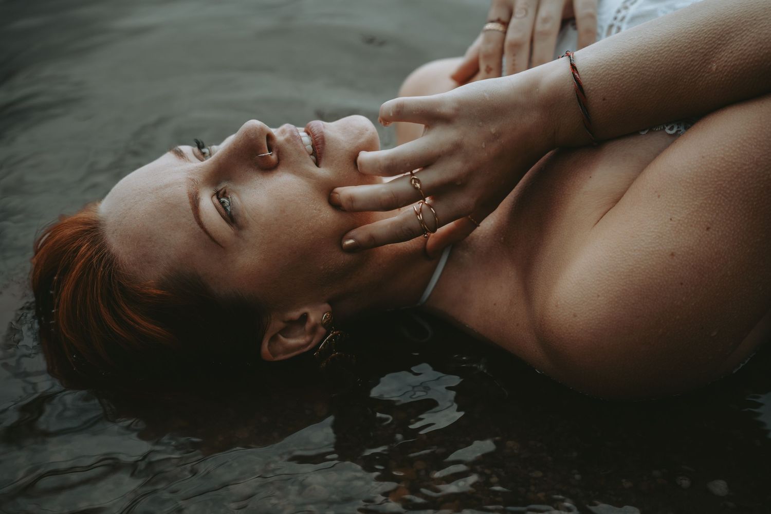 A redheaded woman lies in water, eyes closed, with a hand gently touching her face, wearing gold rings.