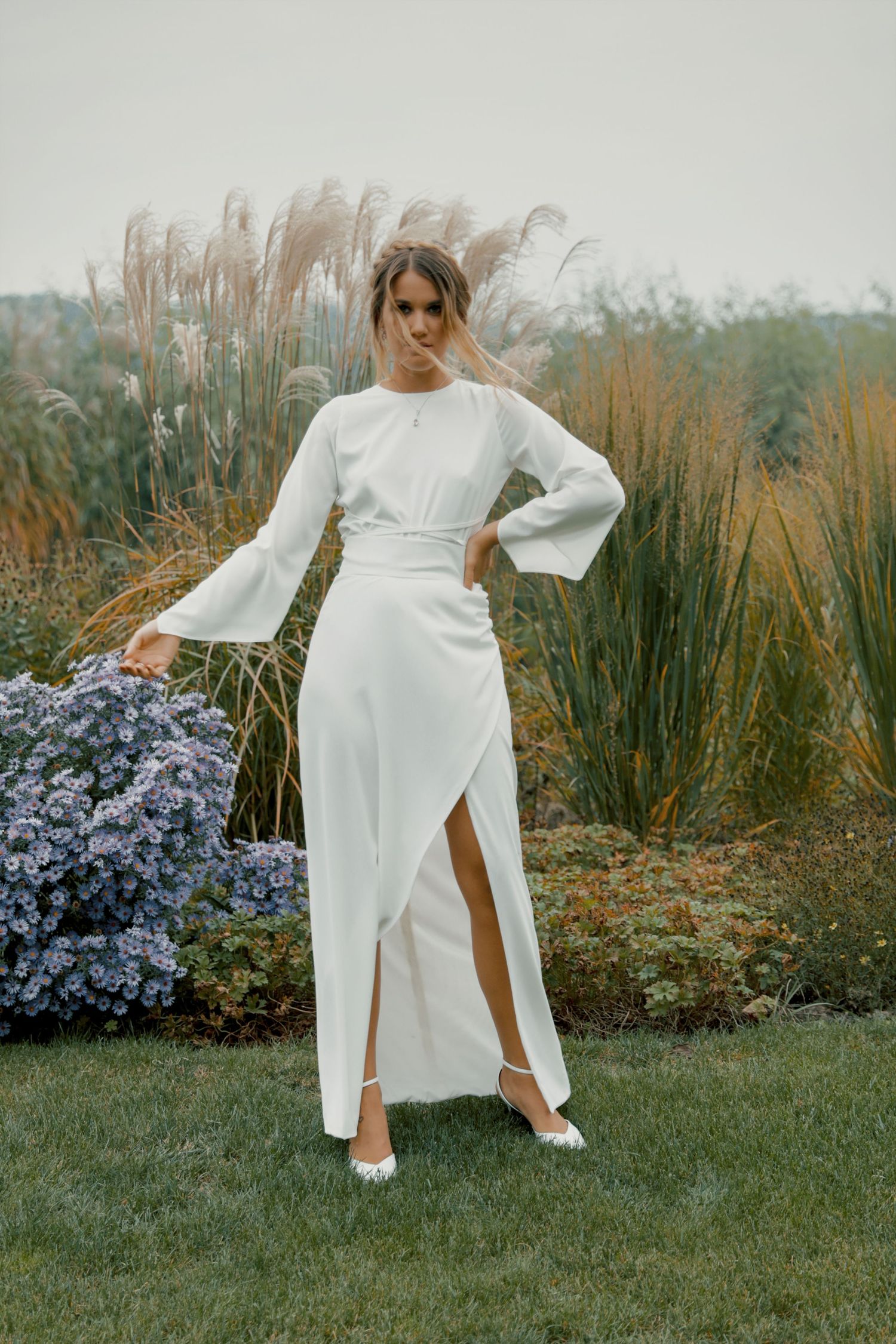 A woman in a white long-sleeve wrap dress poses in a garden with ornamental grasses and flowers.