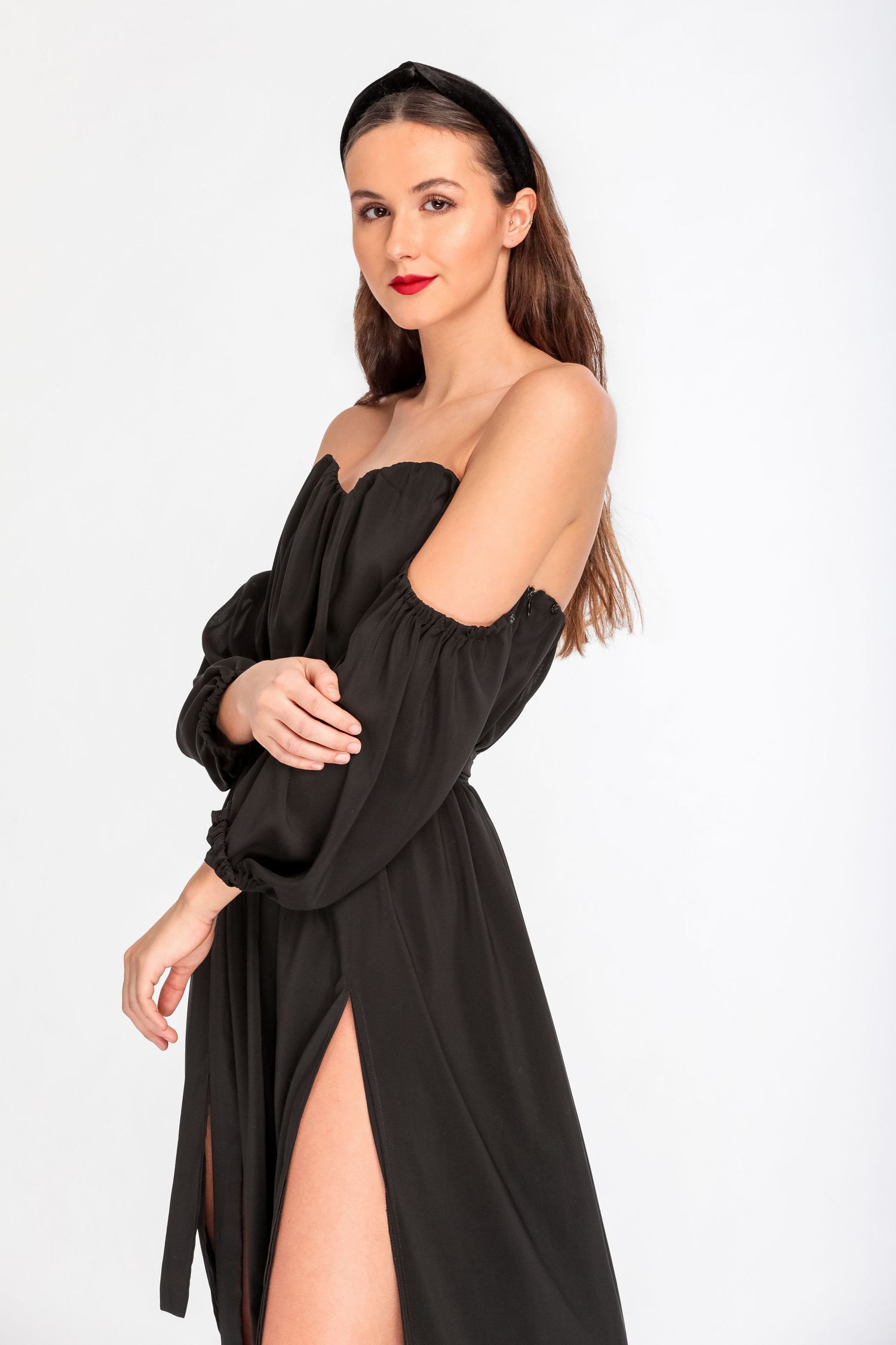 Woman in black off-shoulder maxi dress with long sleeves, side slit, and red lips poses against white background.