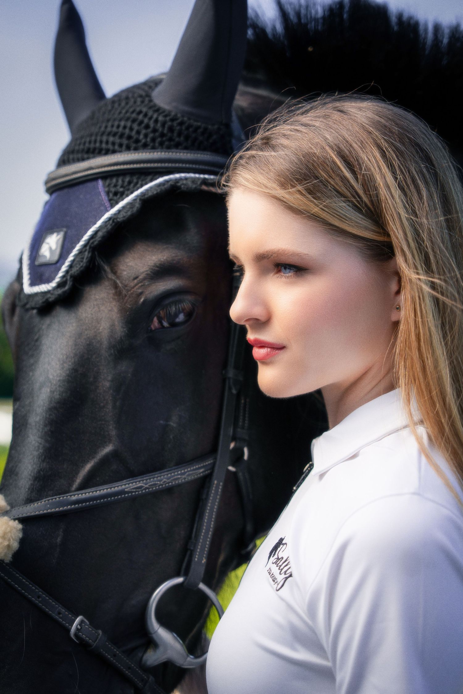 A blonde woman stands close to a black horse wearing a decorative fly bonnet outdoors.