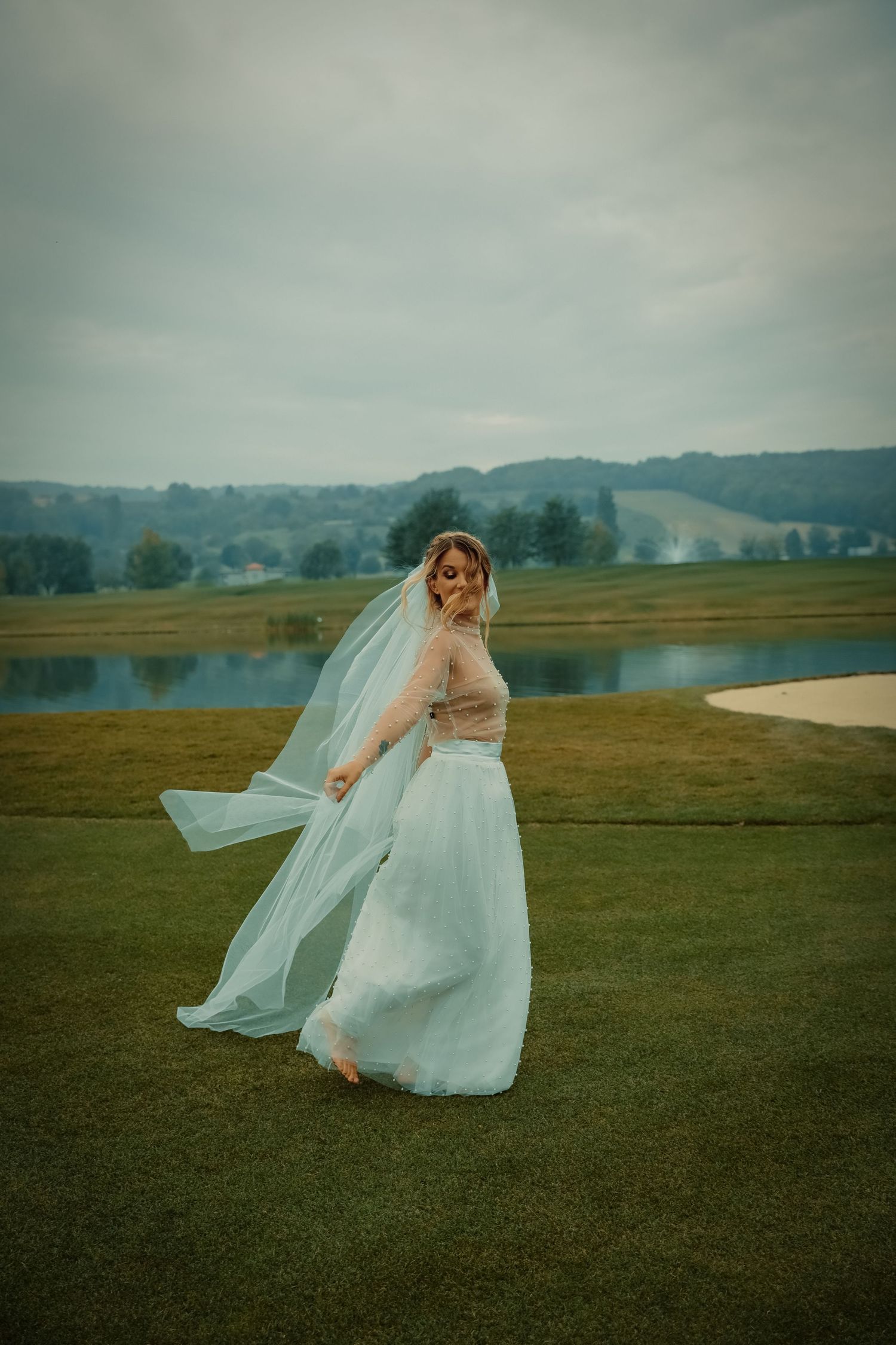 A bride in a flowing light blue skirt and veil twirls on a green golf course near a lake.