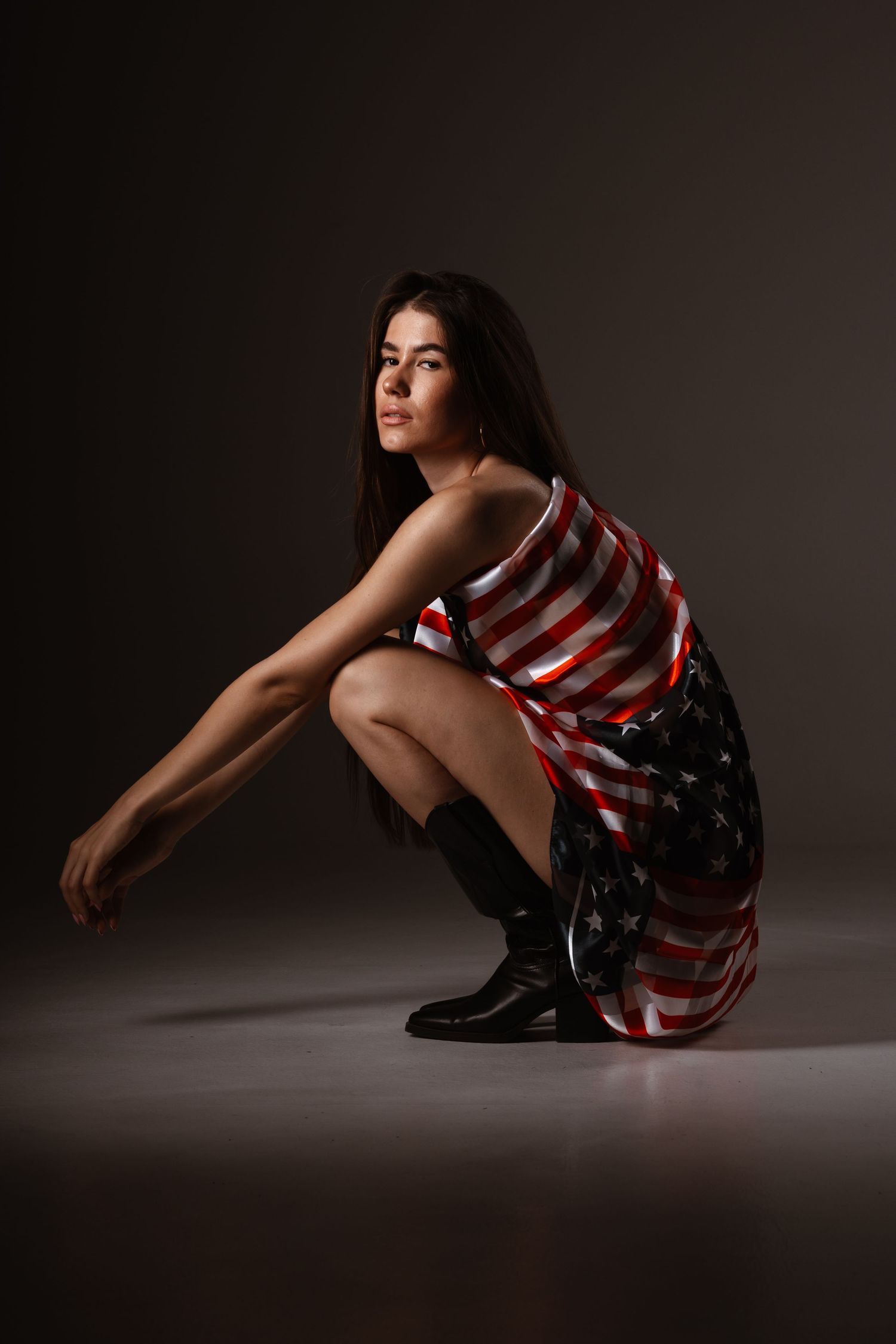 Young woman wearing an American flag dress and black boots, posing seated on a dark studio background.
