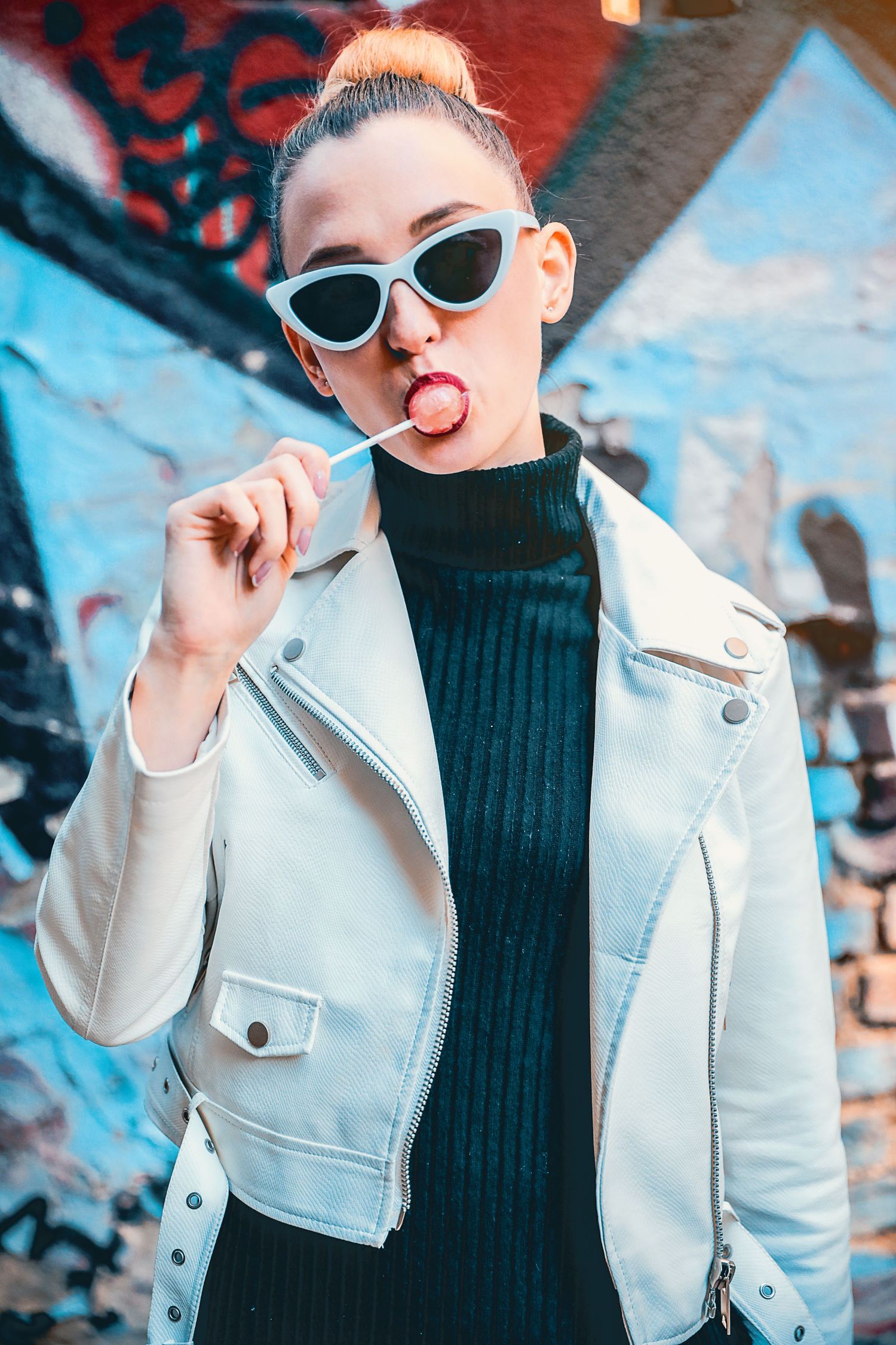 A stylish young woman in a white leather jacket and sunglasses licks a lollipop in front of graffiti.