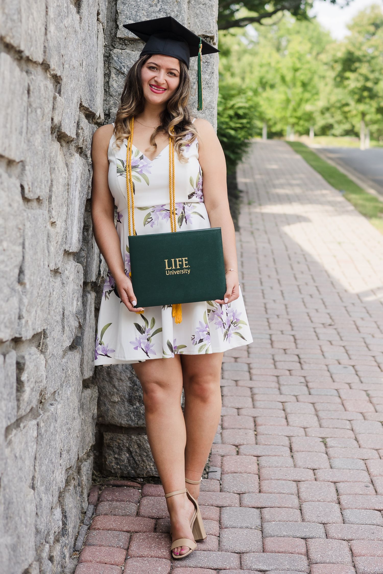 Smiling graduate in floral dress holds Life University diploma while leaning against stone wall.
