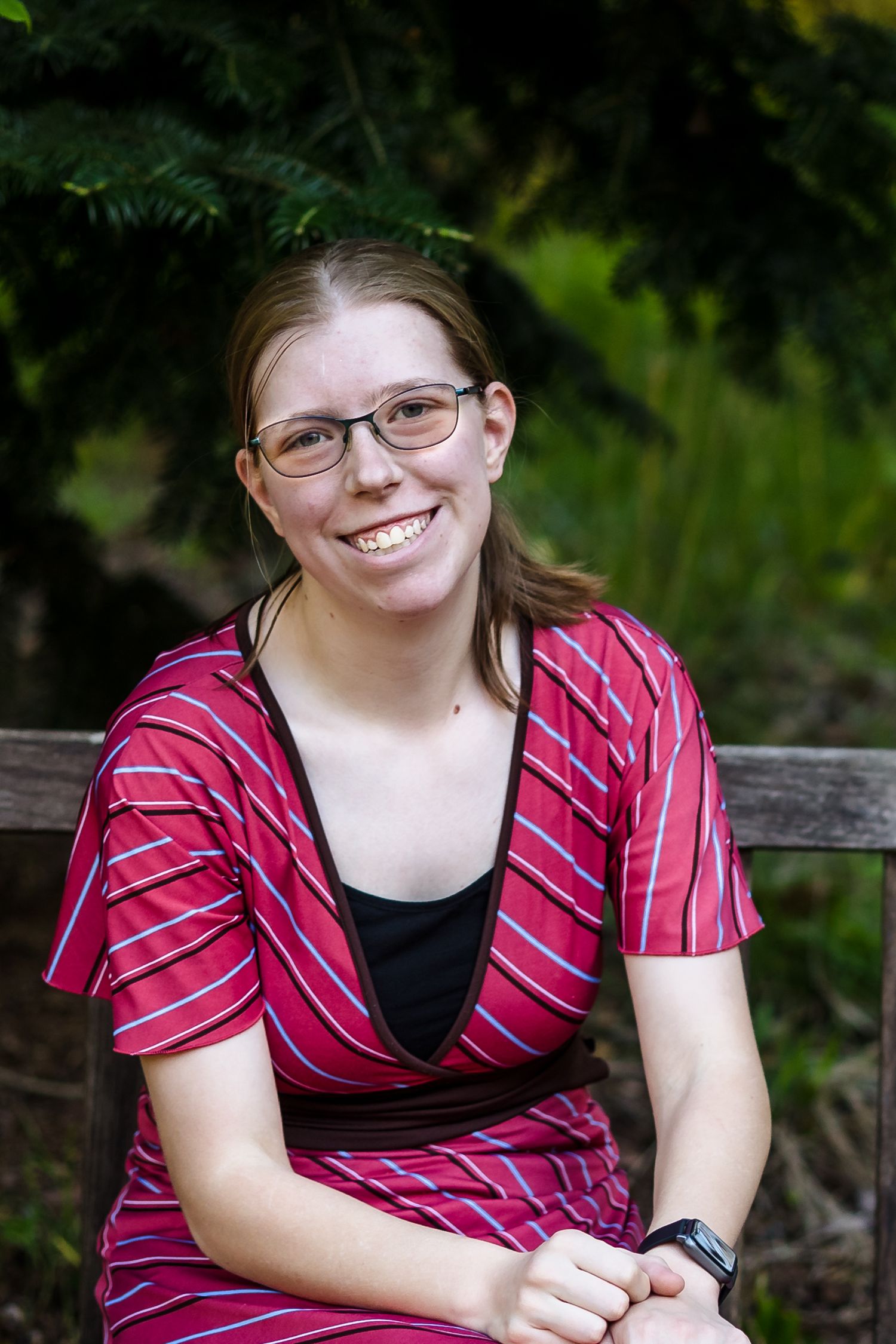 Woman red striped dress portrait — Outdoor portrait of young woman in red striped dress and glasses smiling while seated on a bench.