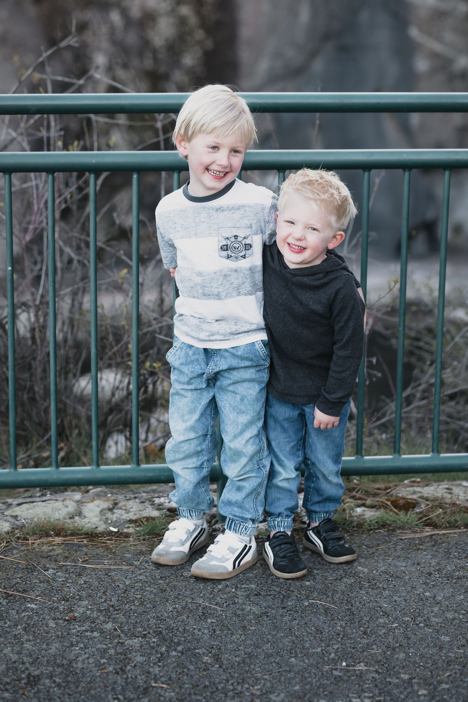 Two blonde boys hugging — Candid outdoor portrait of two young blonde brothers smiling and hugging in front of green metal fence.