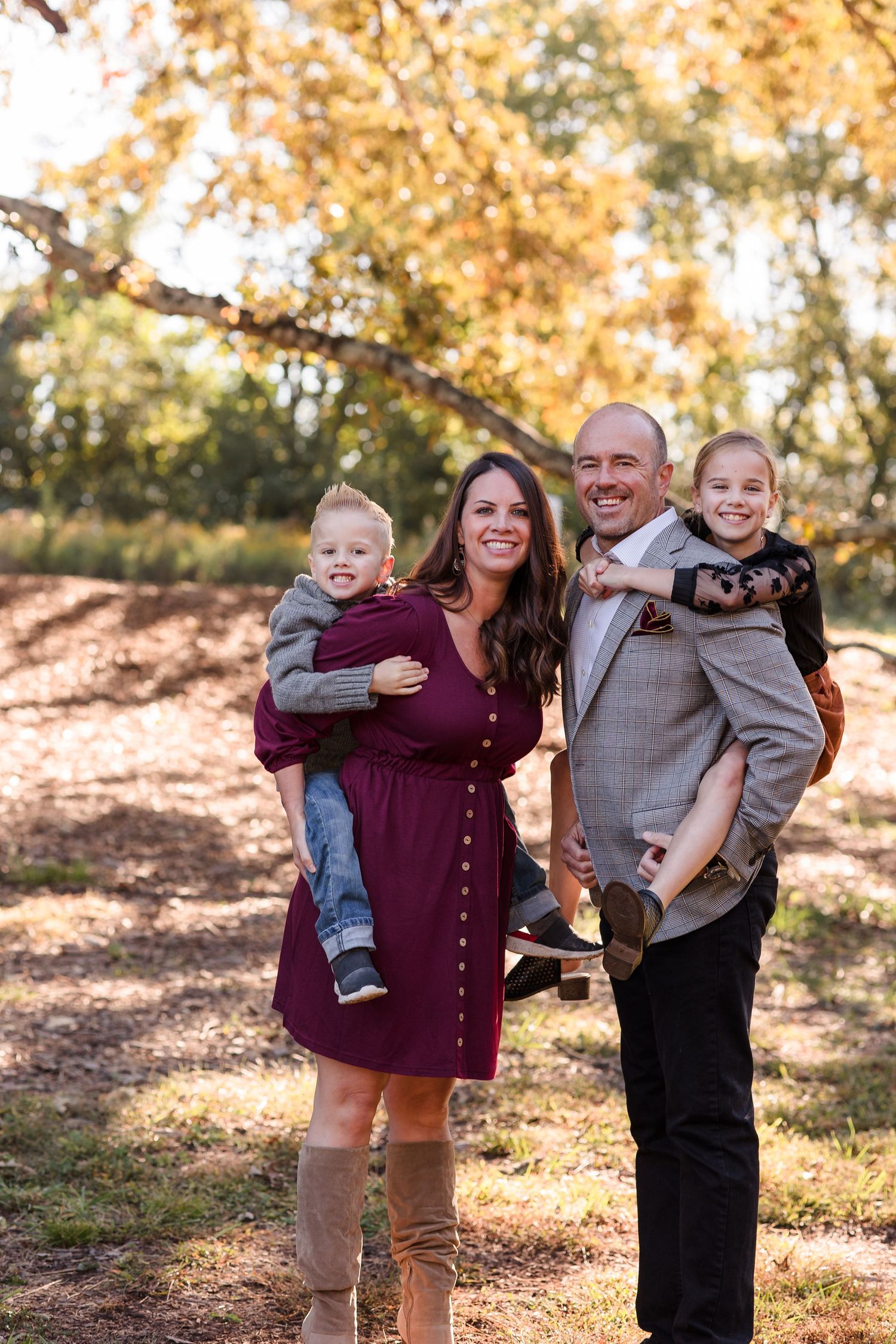 A smiling family of four poses outdoors in fall foliage, parents holding two young children in festive autumn attire.