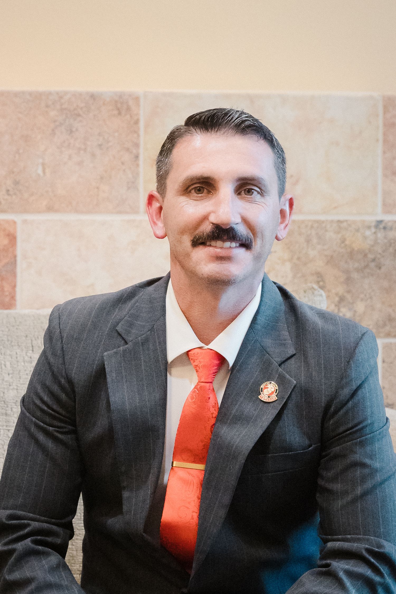 Friendly man with mustache in gray pinstripe suit and orange tie smiling in professional portrait.