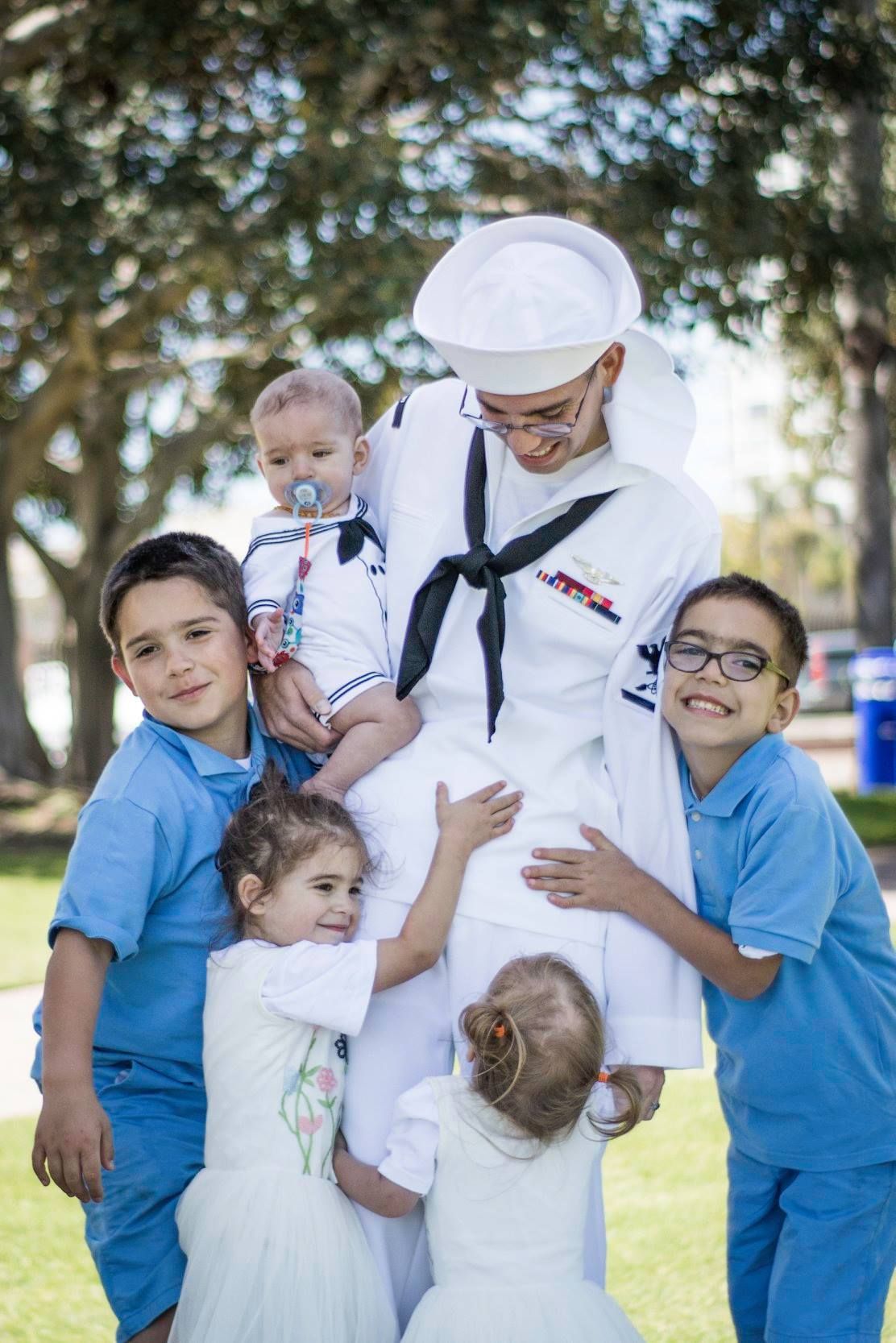 Navy sailor hugged by kids — Navy sailor in white uniform surrounded by five children hugging him during an emotional outdoor moment.