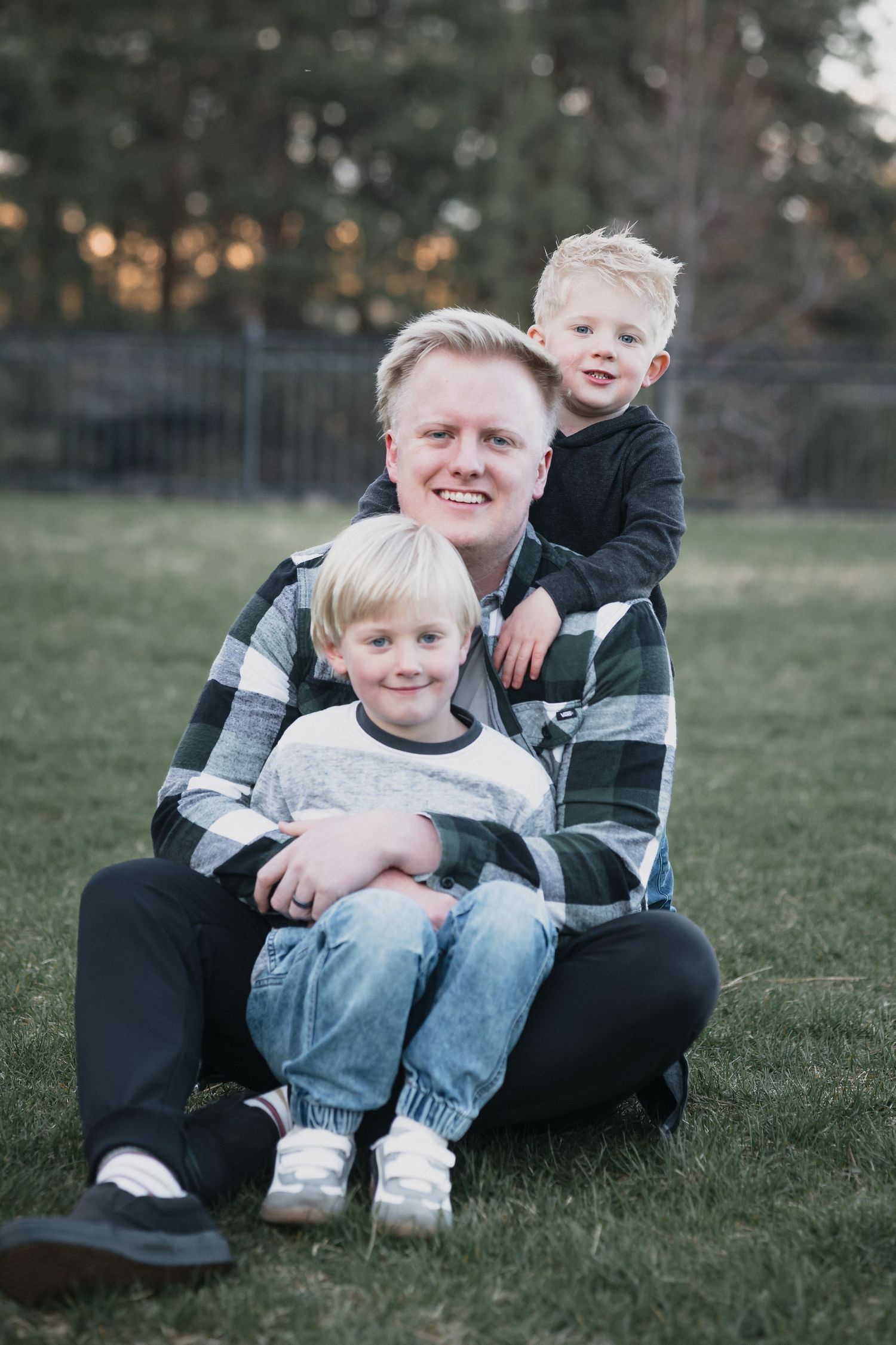 Dad with two sons — Smiling father in a plaid shirt sitting on the grass with his two young blonde sons in a park.