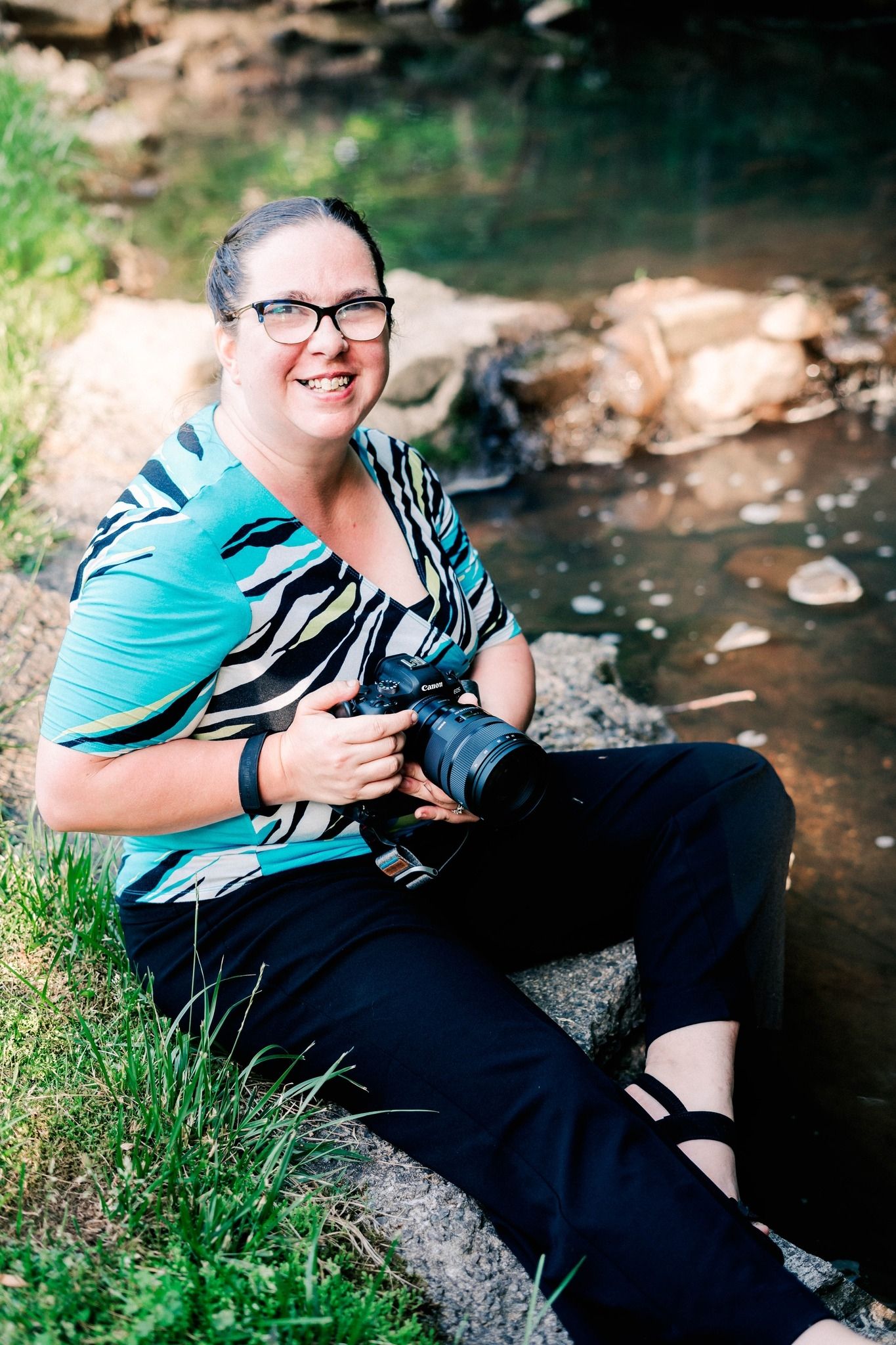 Woman by stream with camera — Woman with glasses sitting by a stream, smiling and holding her camera during an outdoor portrait.