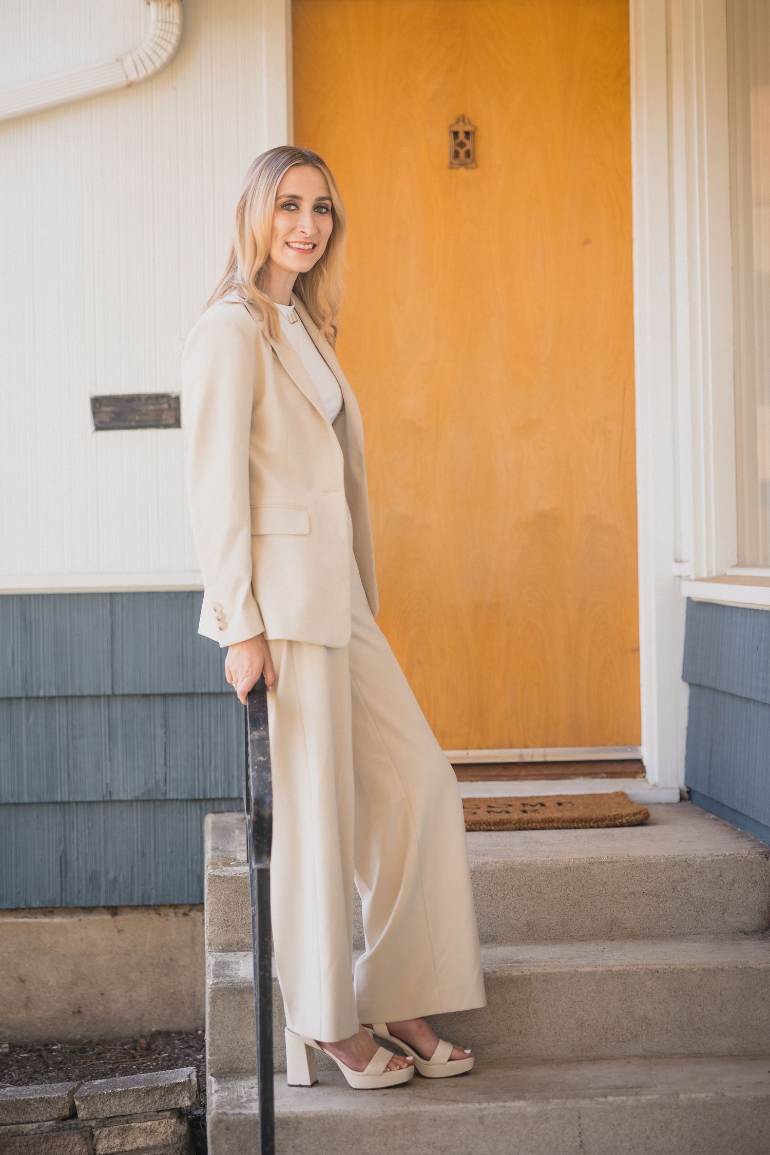 Elegant blonde woman in cream wide-leg pantsuit standing on front porch steps by wooden door.