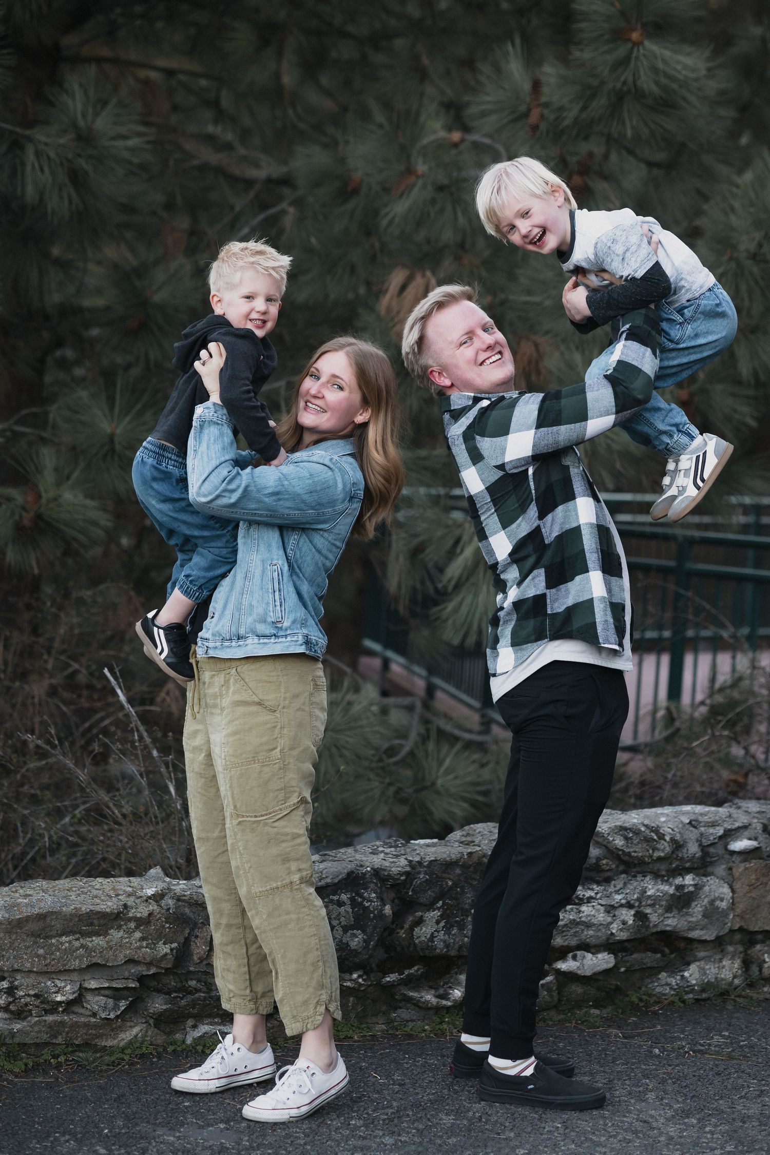 Family lifting two boys — Happy family of four posing outdoors as parents lift their two young blonde boys near a pine tree.