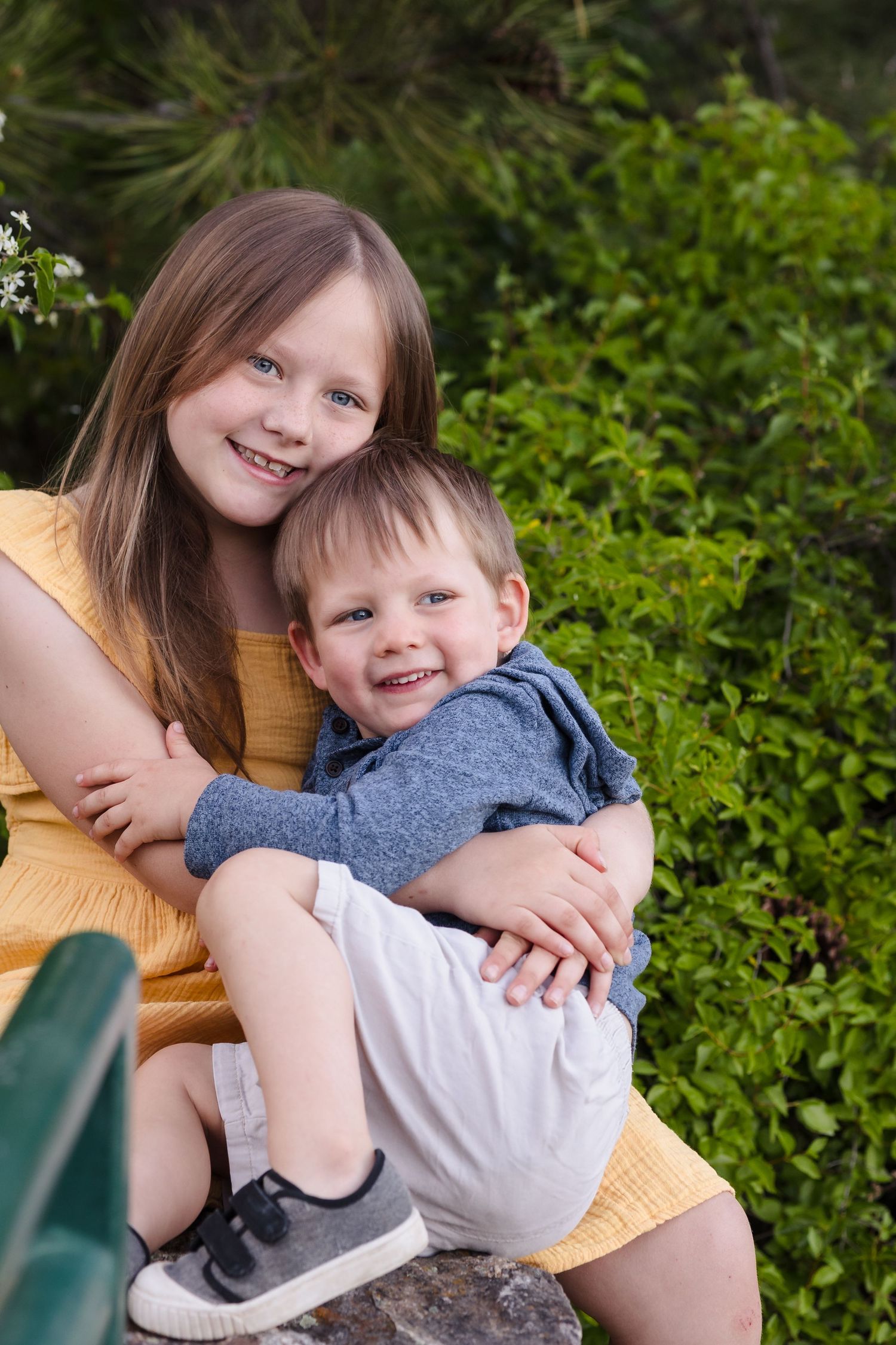 Sister hugging little brother — Smiling older sister in a yellow dress hugging her little brother outdoors near green bushes.