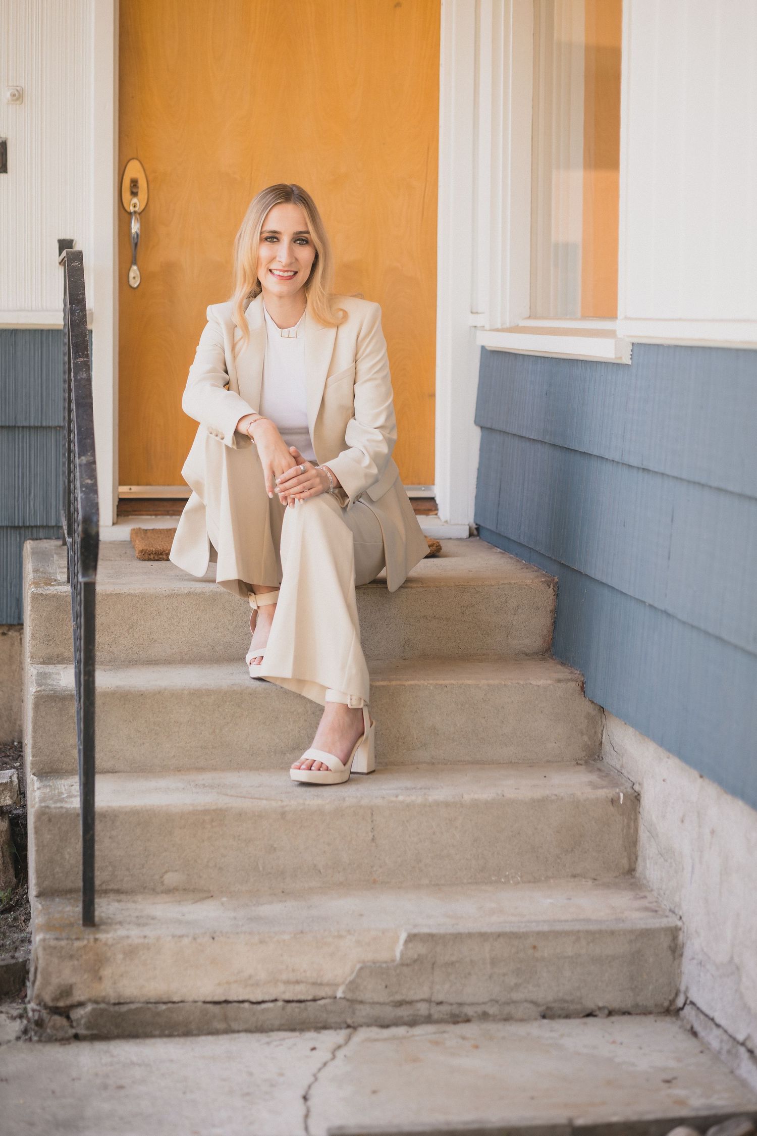 Smiling blonde woman in beige pantsuit sitting on concrete steps outside home with orange door.