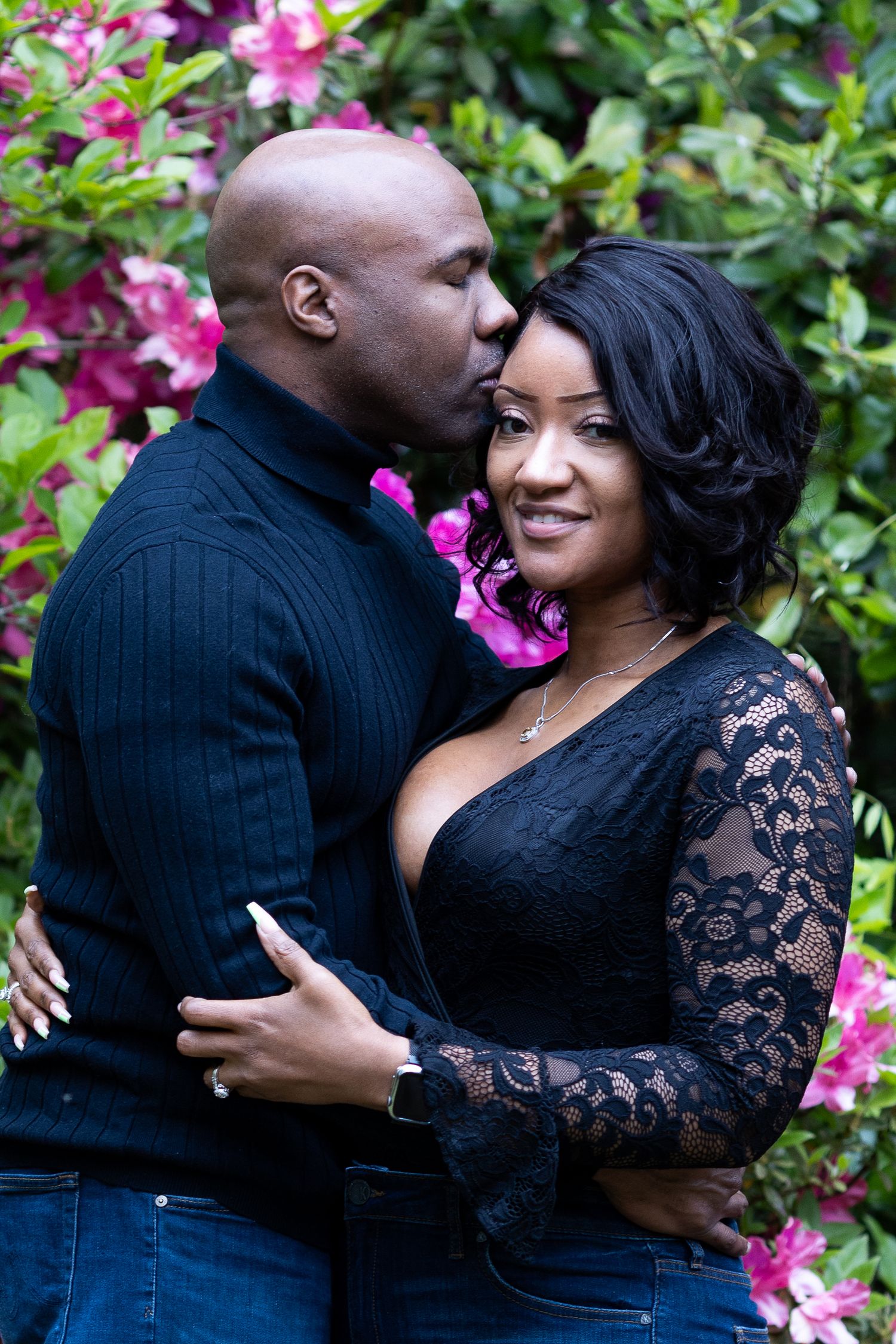Couple by pink flowers — Couple embracing in front of pink flowering bushes, woman in black lace top smiling at the camera.