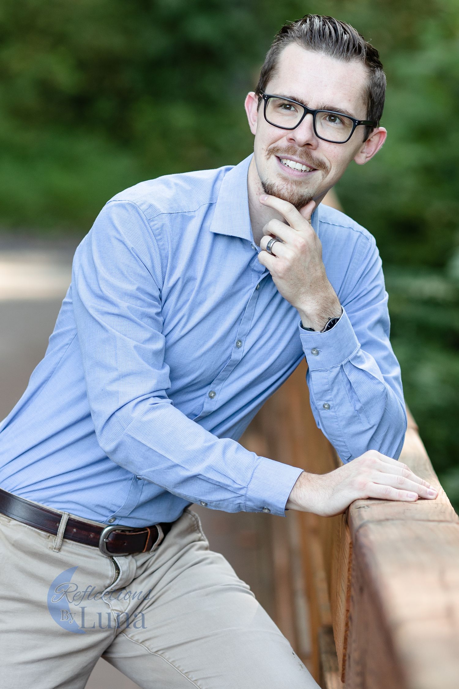 Smiling man in blue shirt and glasses leans on wooden railing outdoors in a relaxed professional headshot.