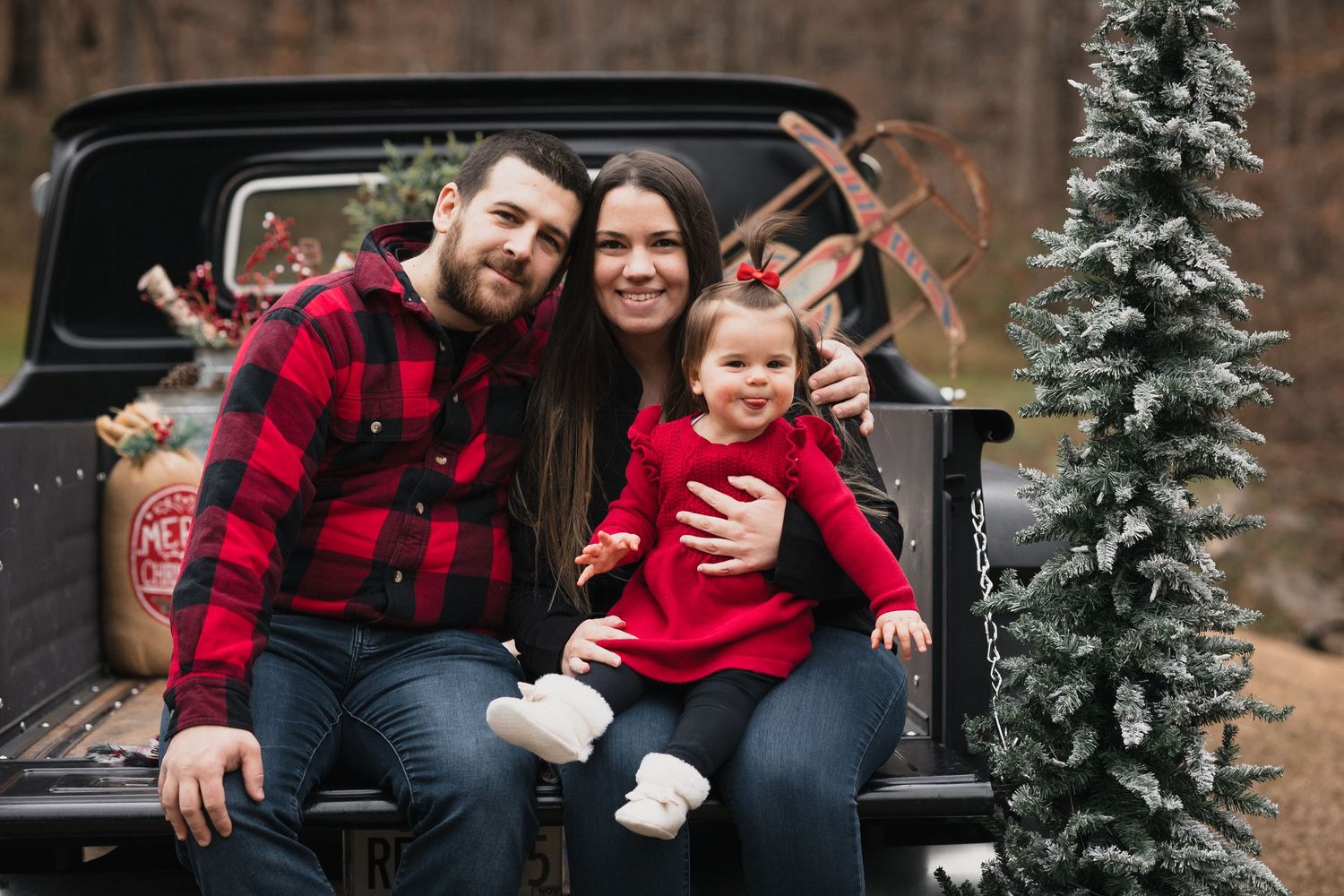 Family of three in red plaid outfits posing on vintage black truck with Christmas trees and reindeer antlers.