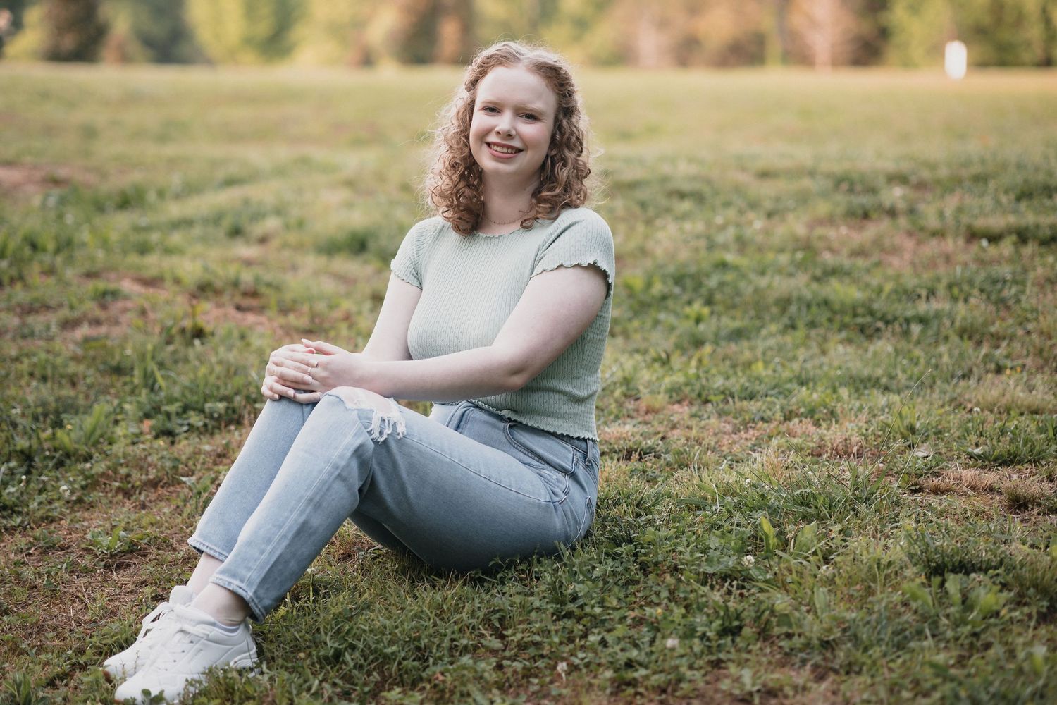 Red headed senior on grass —Curly red hair teen girl sitting on green grass in a park, wearing a mint shirt and light jeans, smiling warmly.