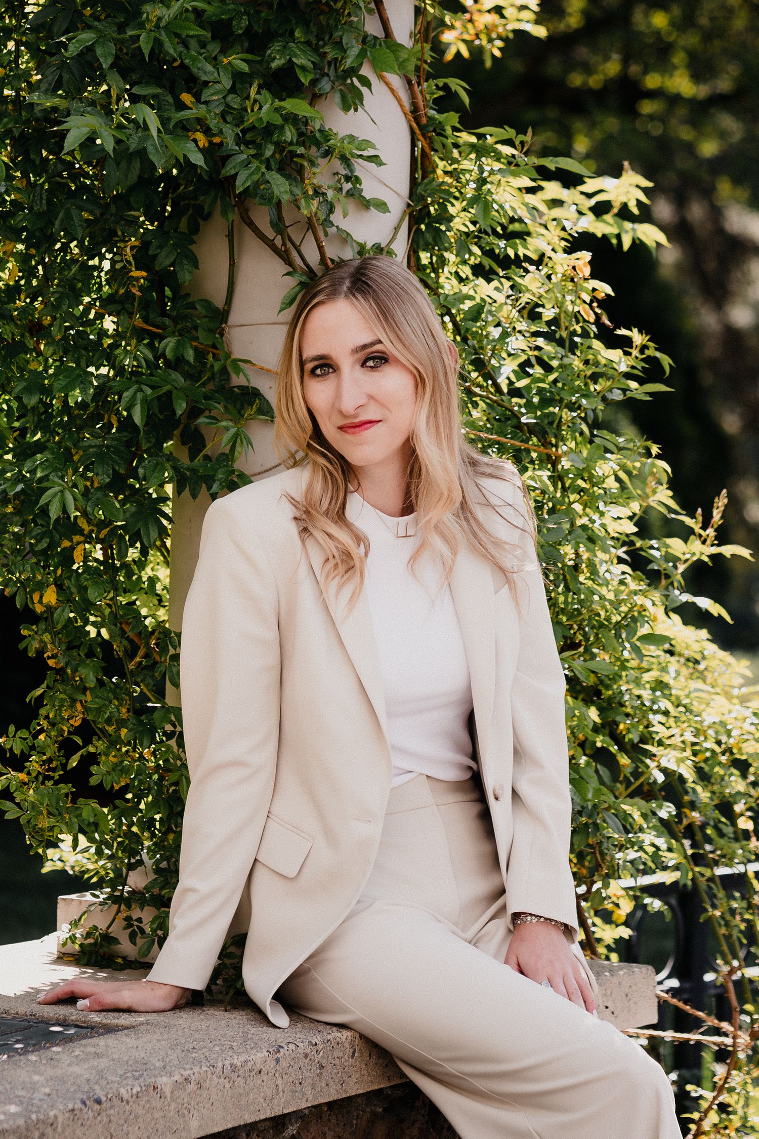Woman in beige blazer suit sitting outdoors against lush green ivy-covered wall in elegant professional portrait.