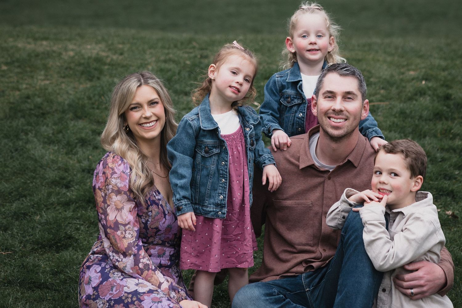 Family of five on grass — Outdoor family portrait of parents and three young children smiling together on green grass in warm, natural light
